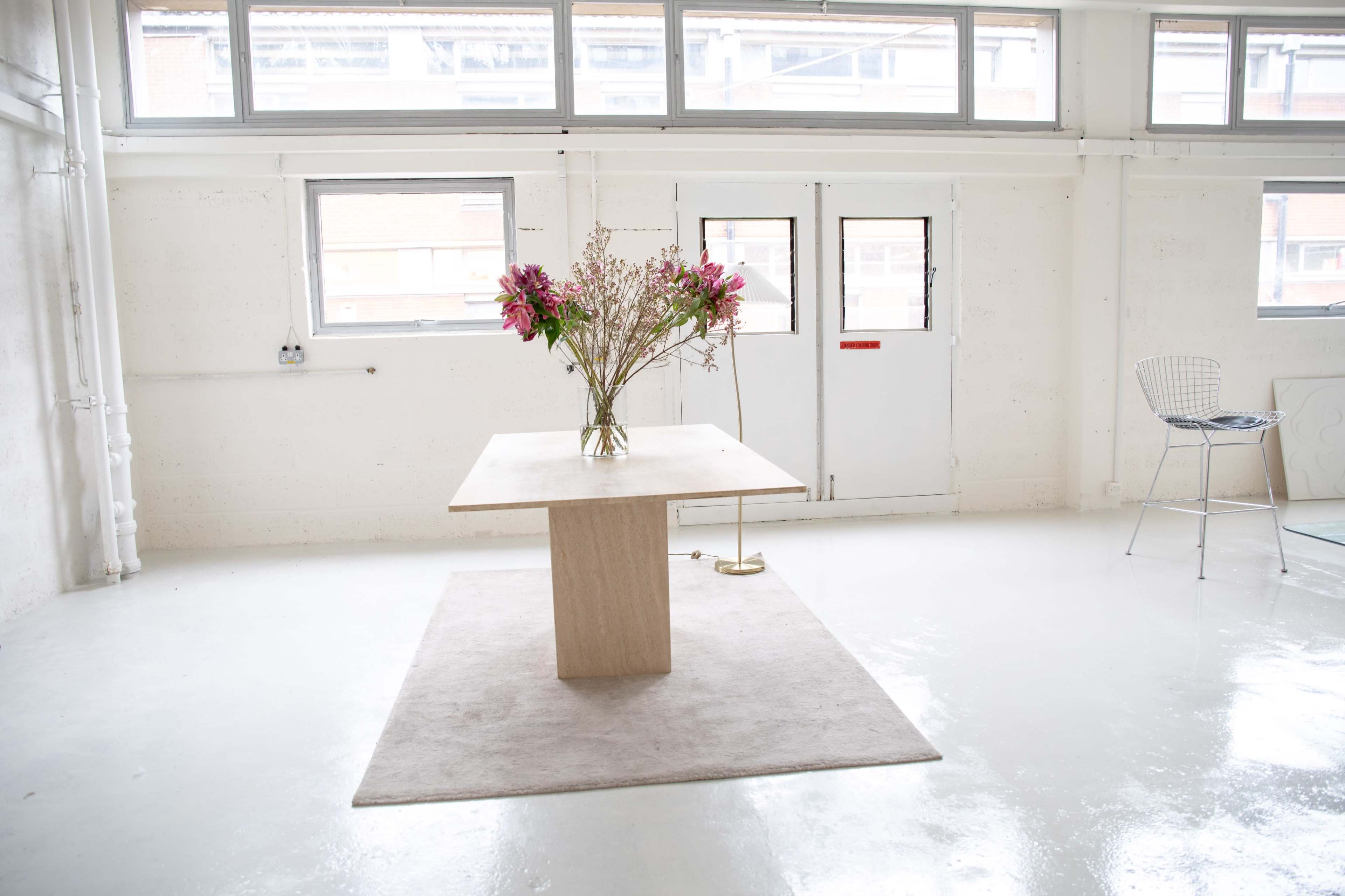 A minimalist room with white walls and floor features a central table with a vase of flowers, a small rug underneath, and a chair nearby.