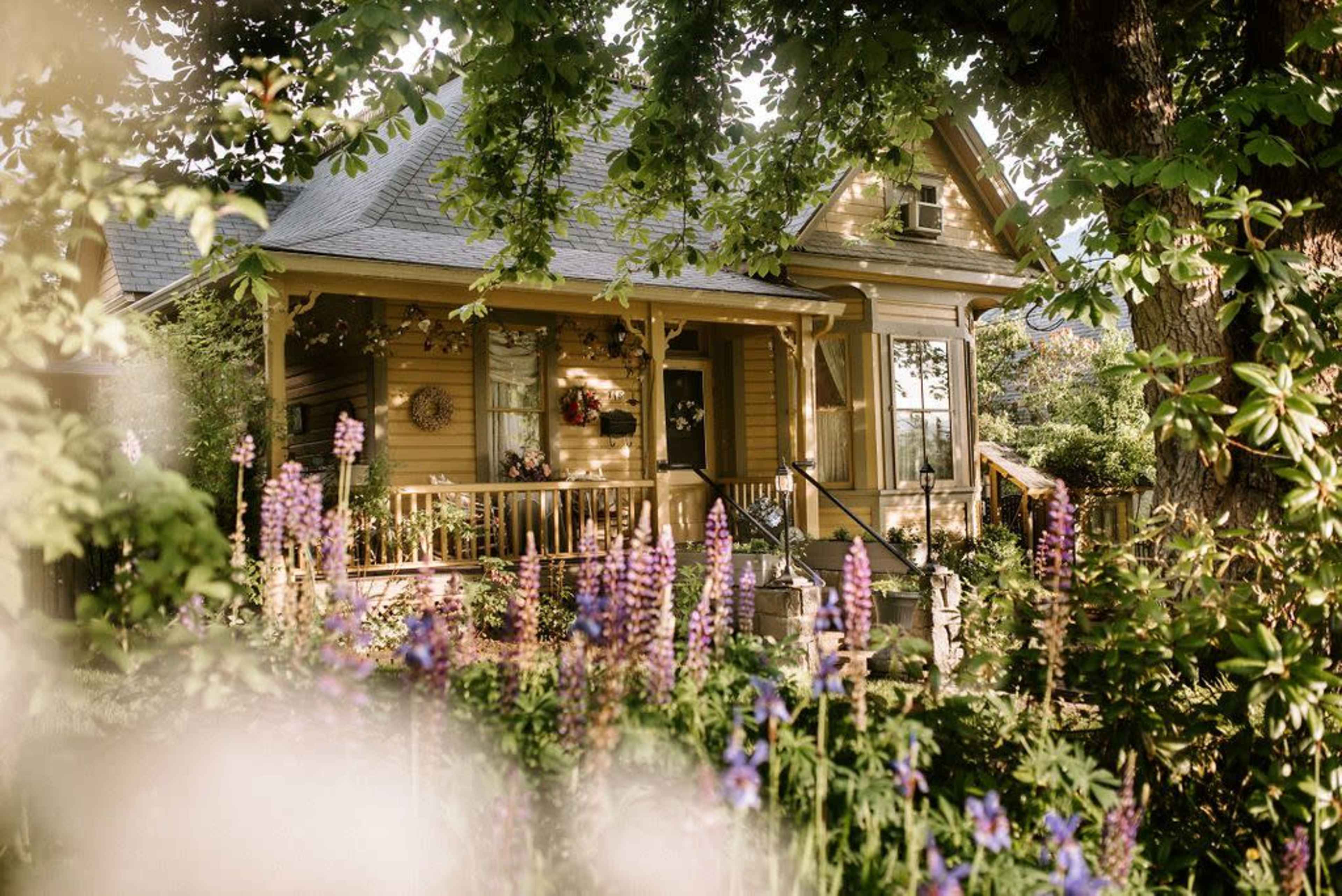 A yellow Victorian-style house stands surrounded by colorful flowers and greenery.