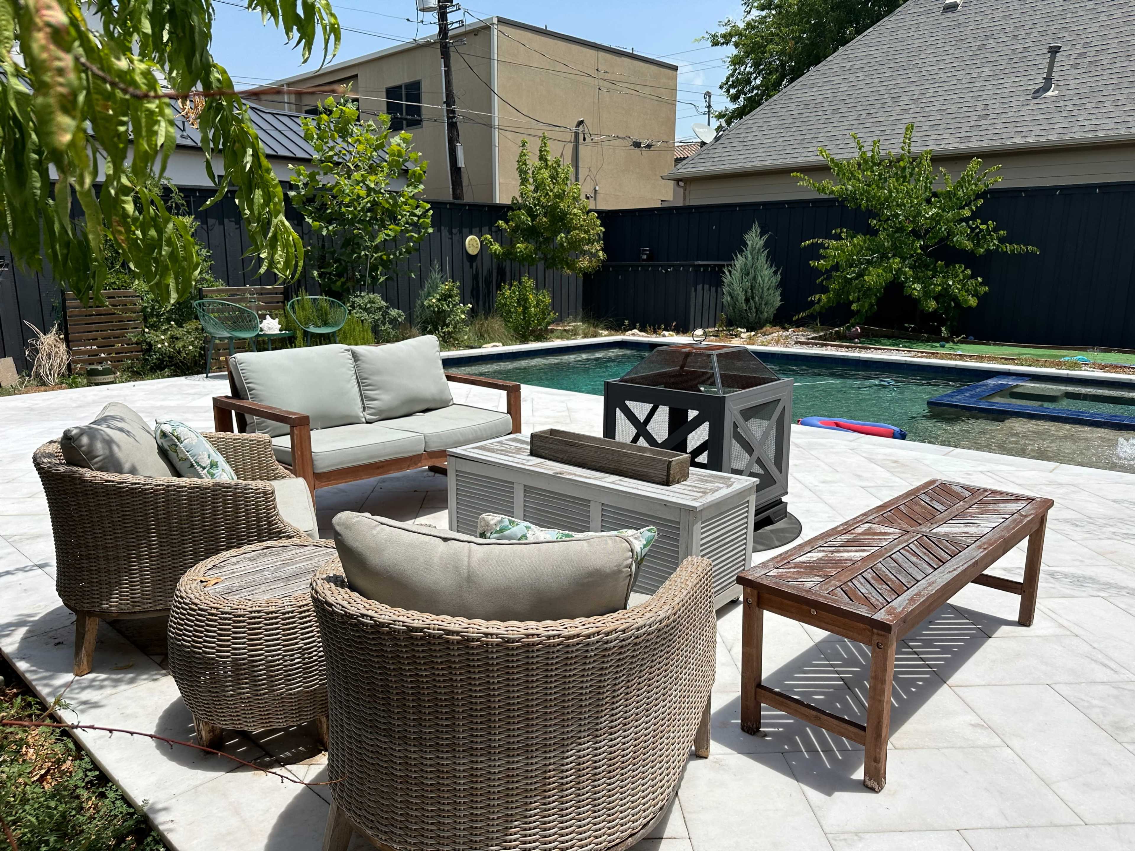 A patio area with a seating arrangement of wicker chairs and a coffee table, positioned around a swimming pool surrounded by greenery and fences.