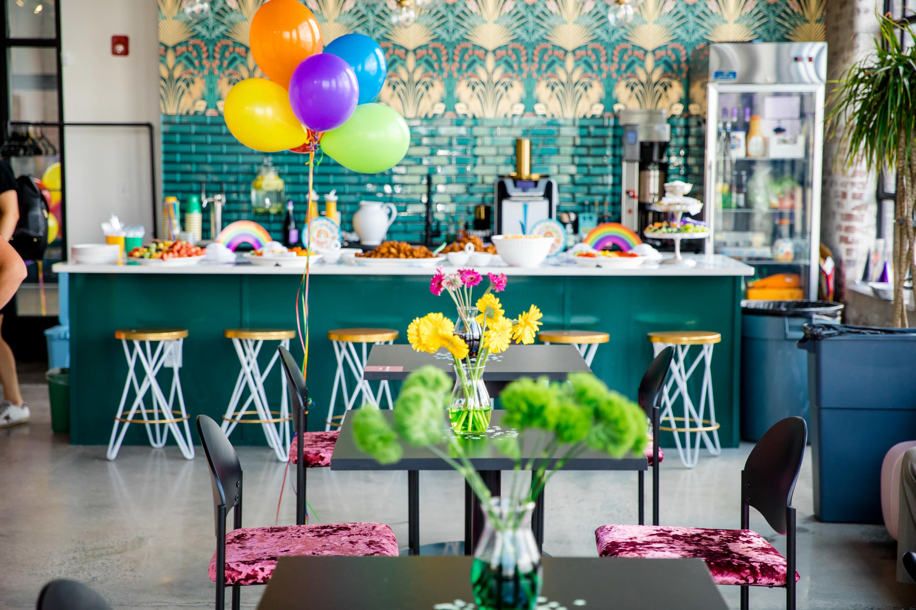 A brightly decorated café with colorful balloons, flowers on tables, and a vibrant green counter filled with food.