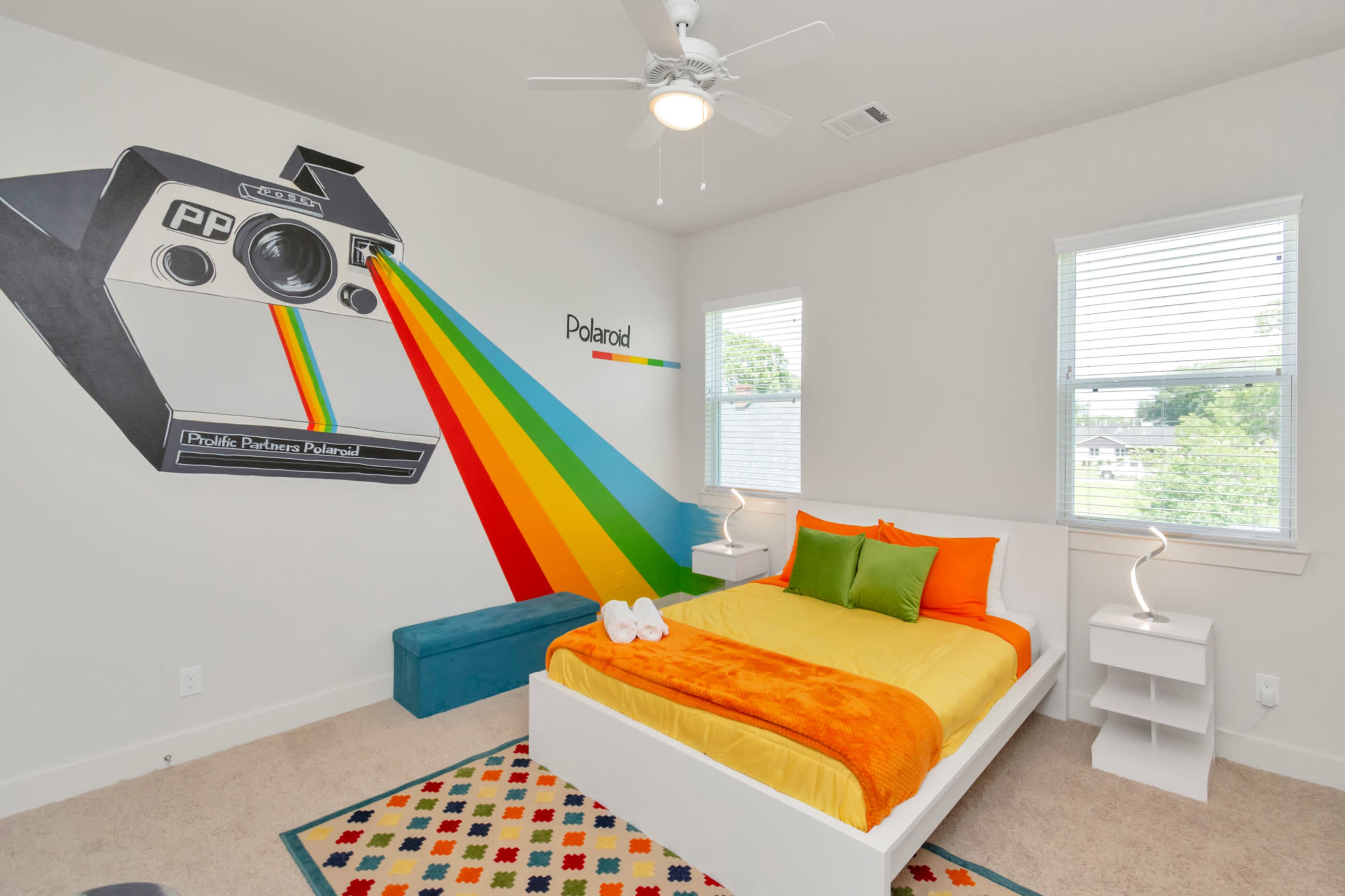 A bright bedroom featuring a large Polaroid camera mural on the wall, a bed with yellow and green bedding, and a colorful rug on the carpeted floor.