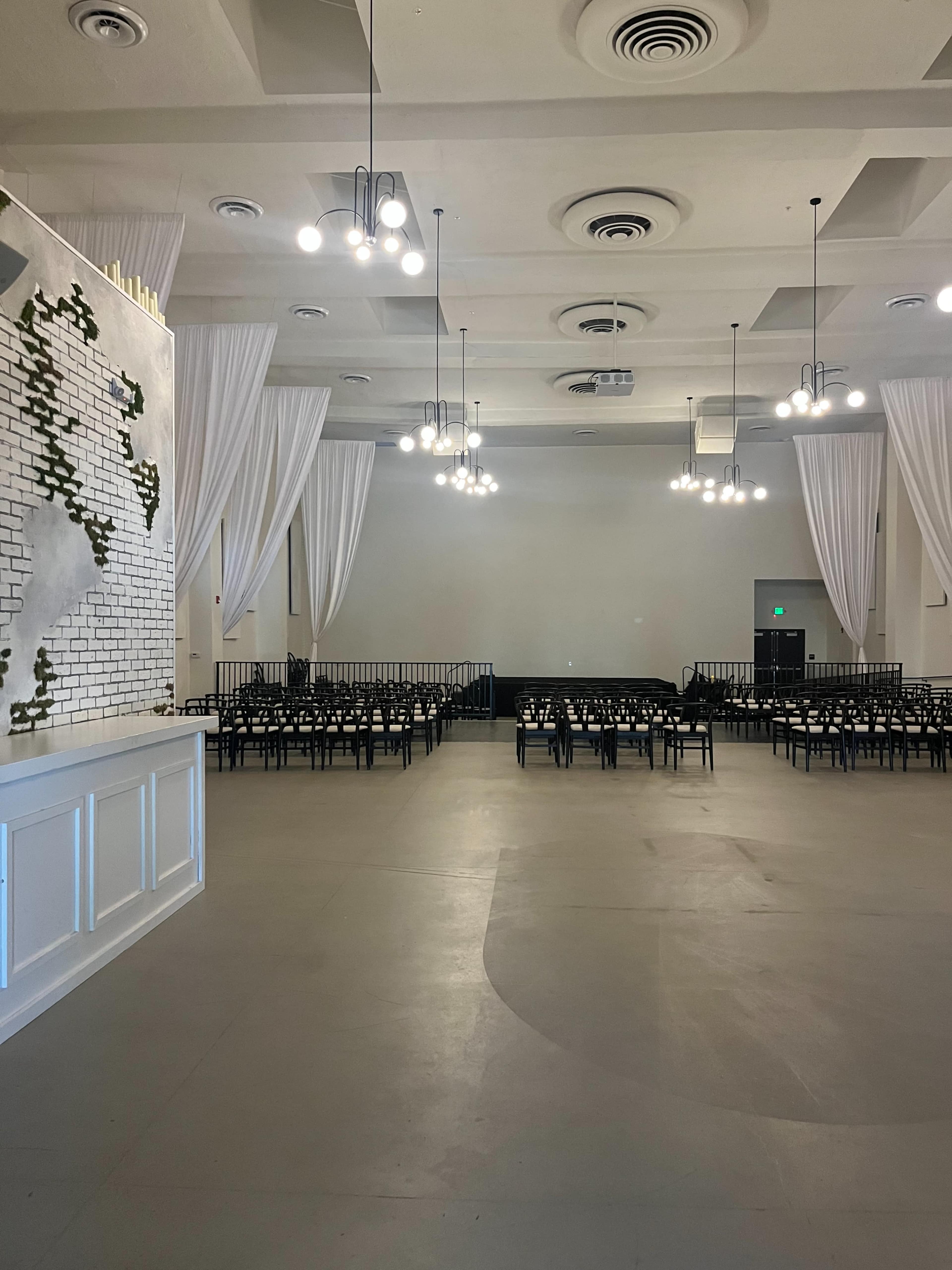 A spacious event venue with white drapes, pendant lights, and rows of black chairs arranged in front of a blank wall.