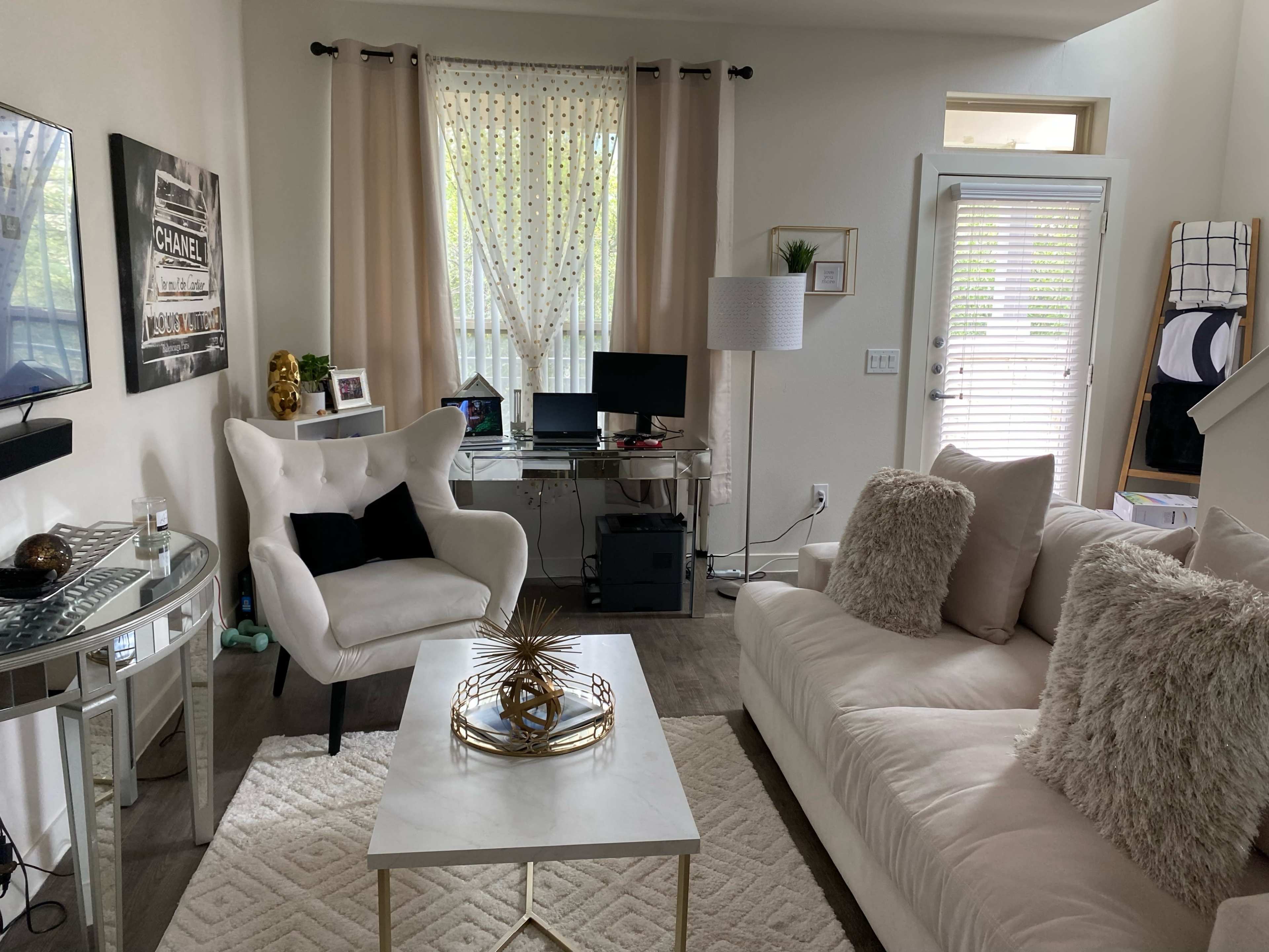 The image shows a living room with a white sofa, a glass coffee table, a computer desk, and large windows covered with light curtains.