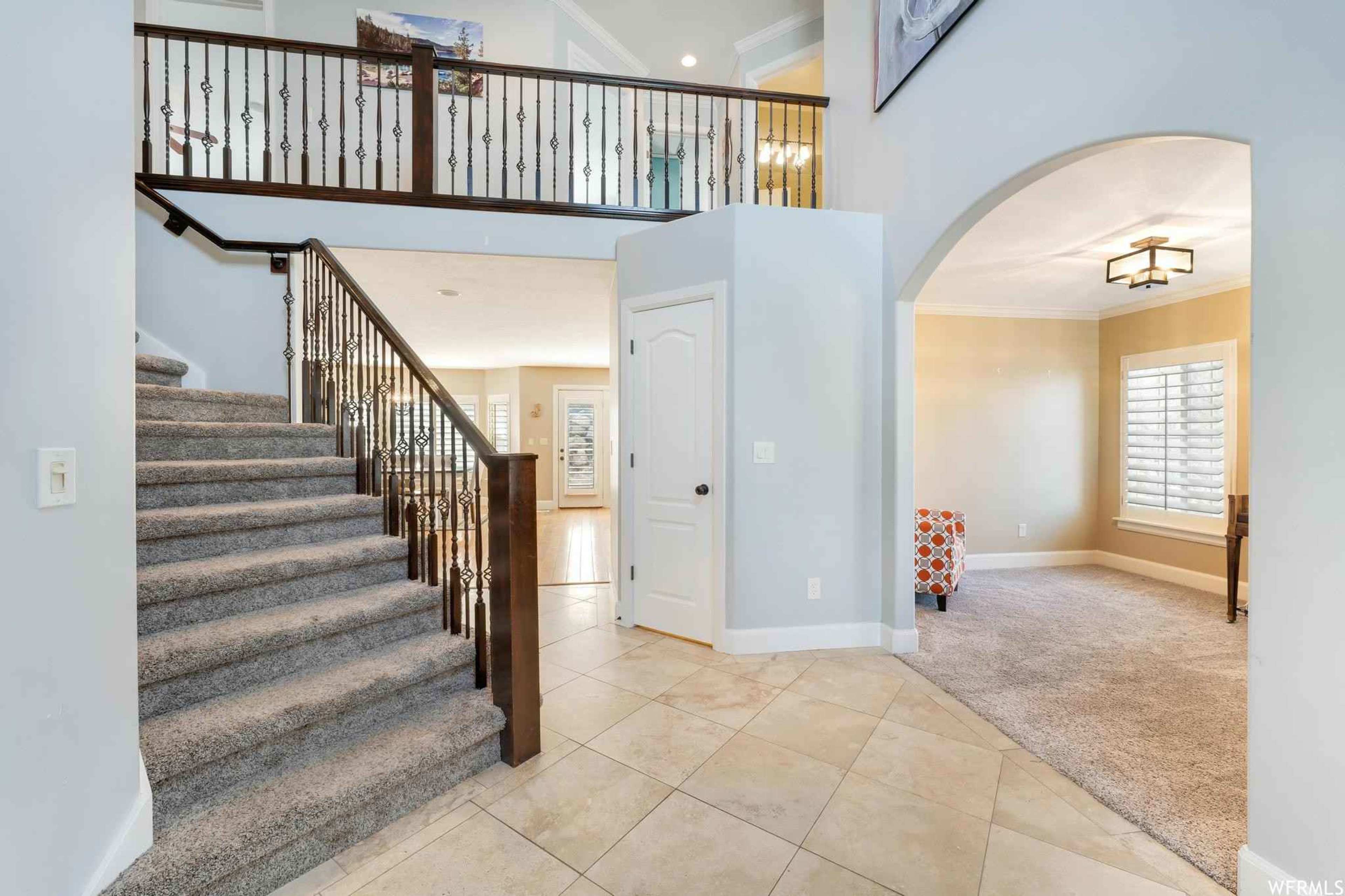 A staircase leads from a tiled entryway to a second floor, framed by a decorative railing, with an archway opening to a carpeted living area on the right.