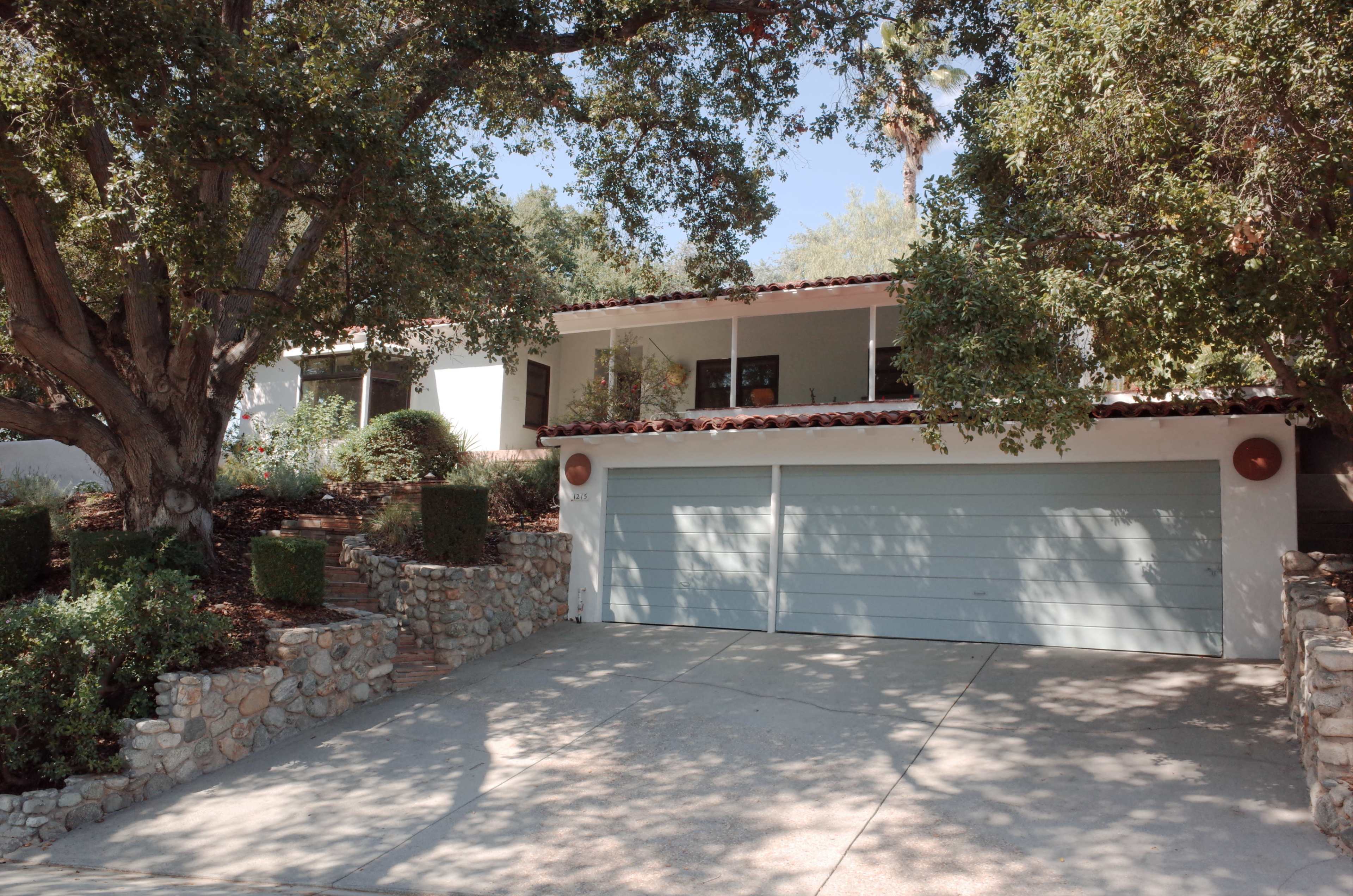 A house with a two-car garage, surrounded by greenery and situated on a sloped driveway.