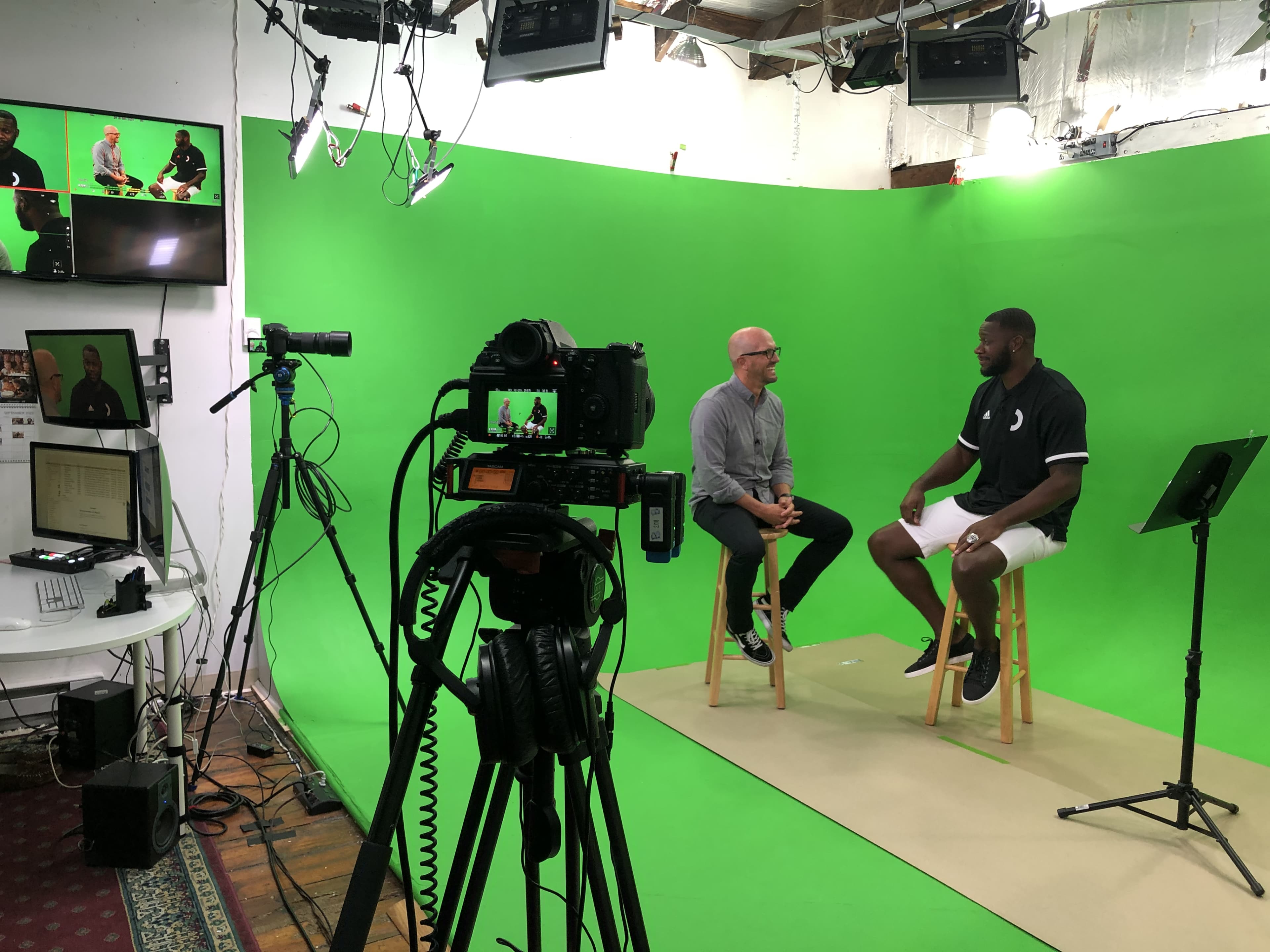 A two-person interview is taking place in front of a green screen, with cameras and lighting equipment set up in a studio.