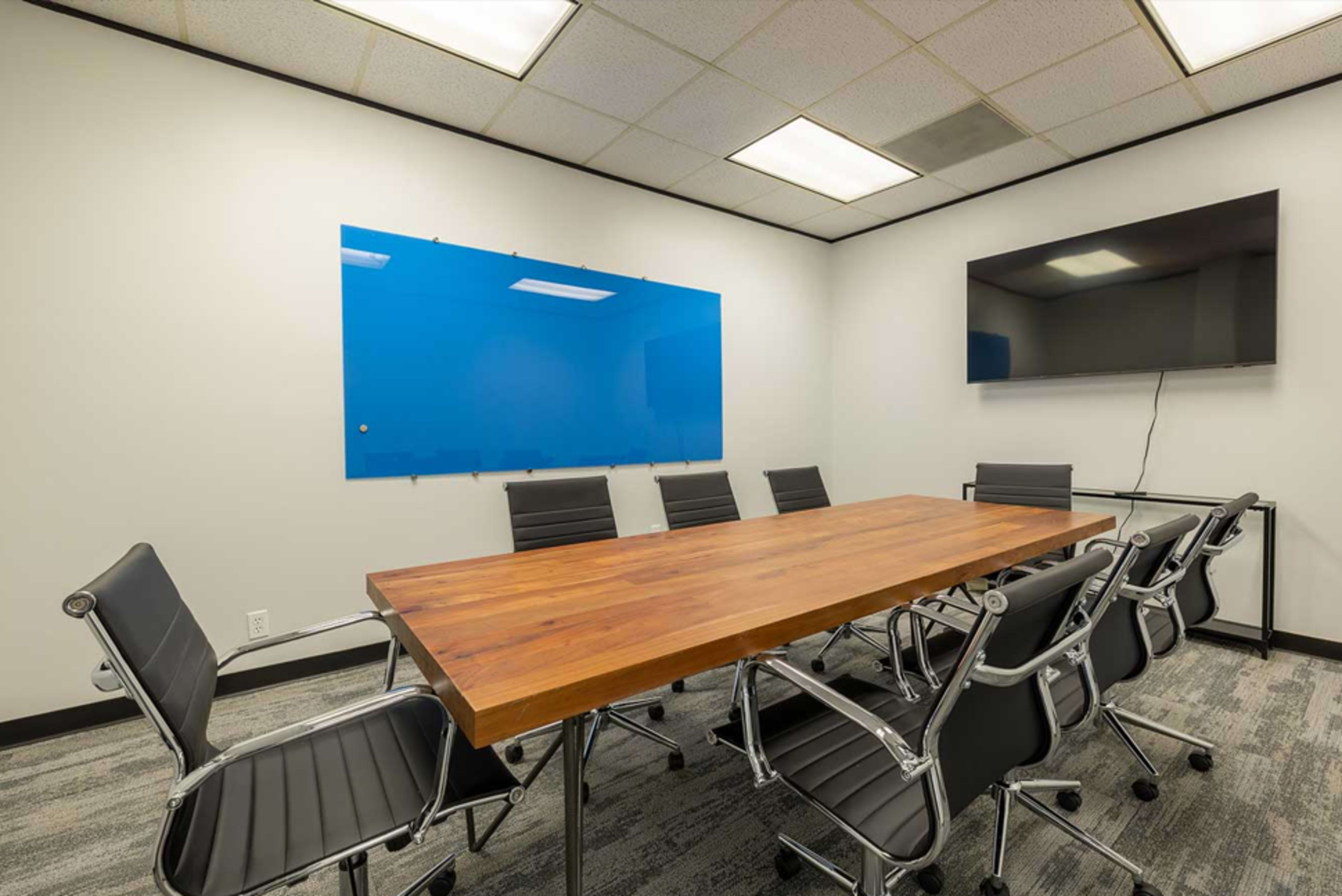 The image shows a modern conference room with a long wooden table surrounded by black office chairs, a blue whiteboard on the wall, and a wall-mounted television.
