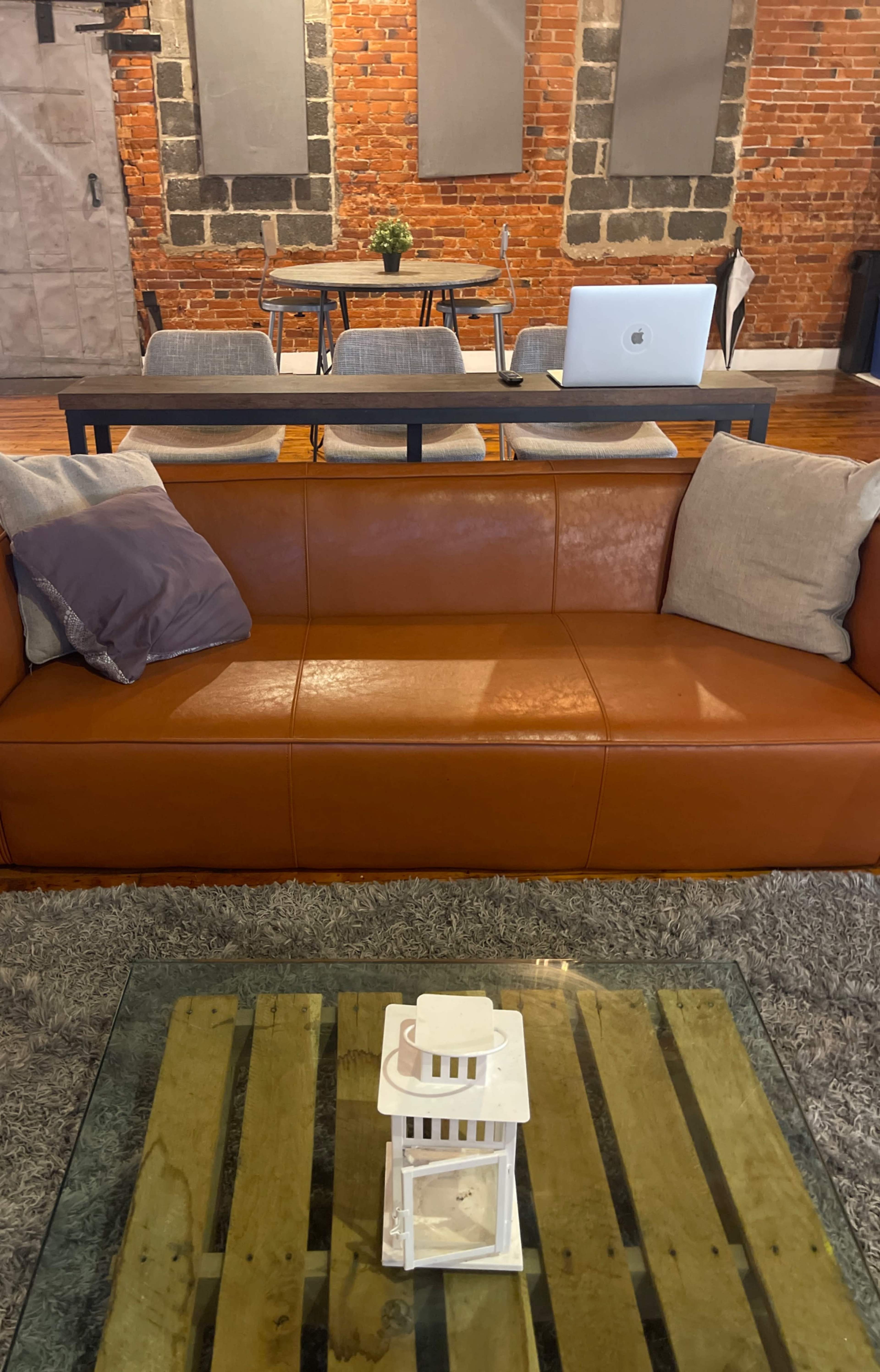 The image features a brown leather couch with gray cushions in a room that has exposed brick walls and a wooden table with a laptop on it.