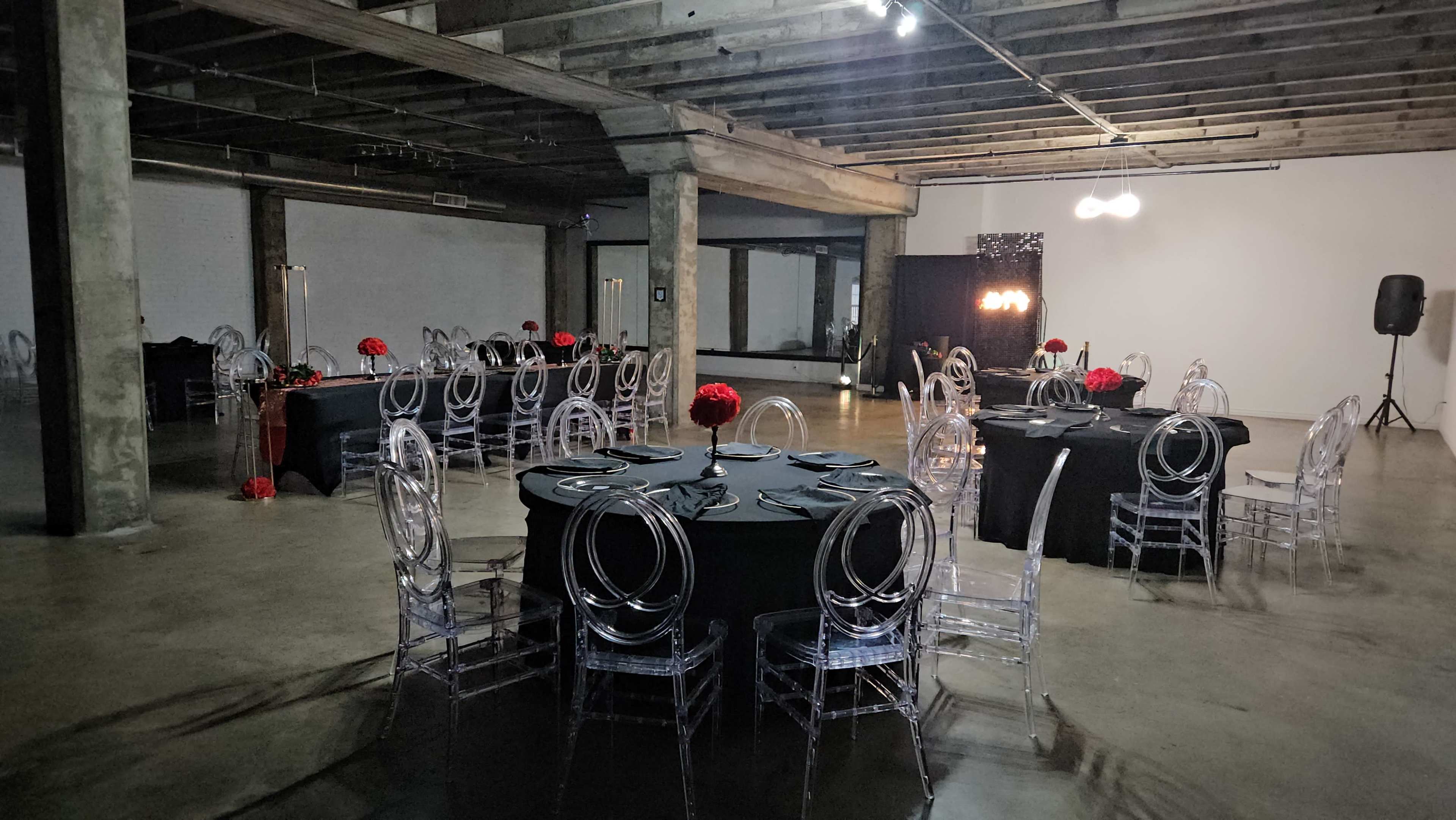 The image shows an industrial-style event space set with black tables and transparent chairs, adorned with red floral centerpieces.