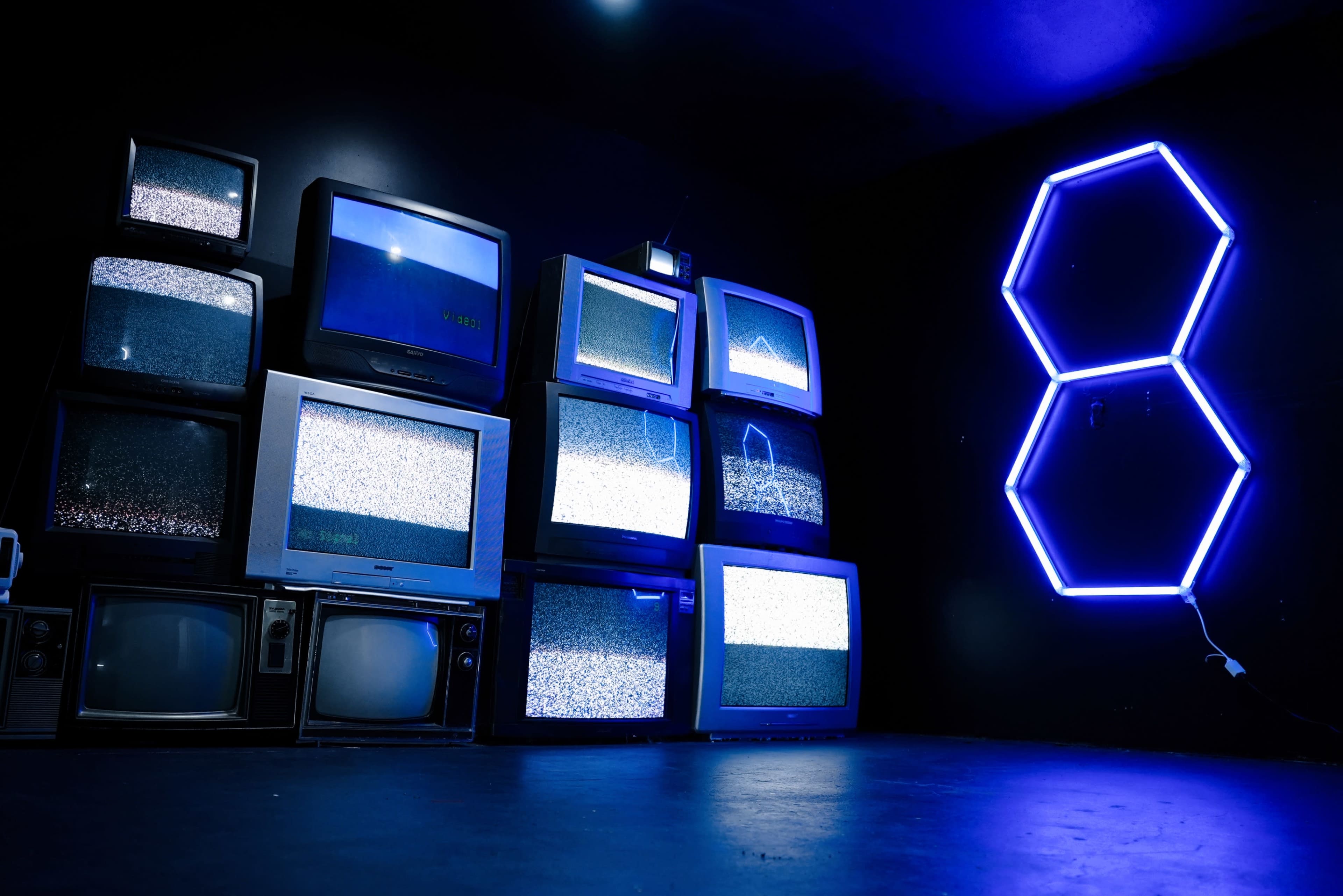 The image shows a stack of vintage television sets displaying static on their screens against a dark background, with a neon blue number eight illuminated on the wall.