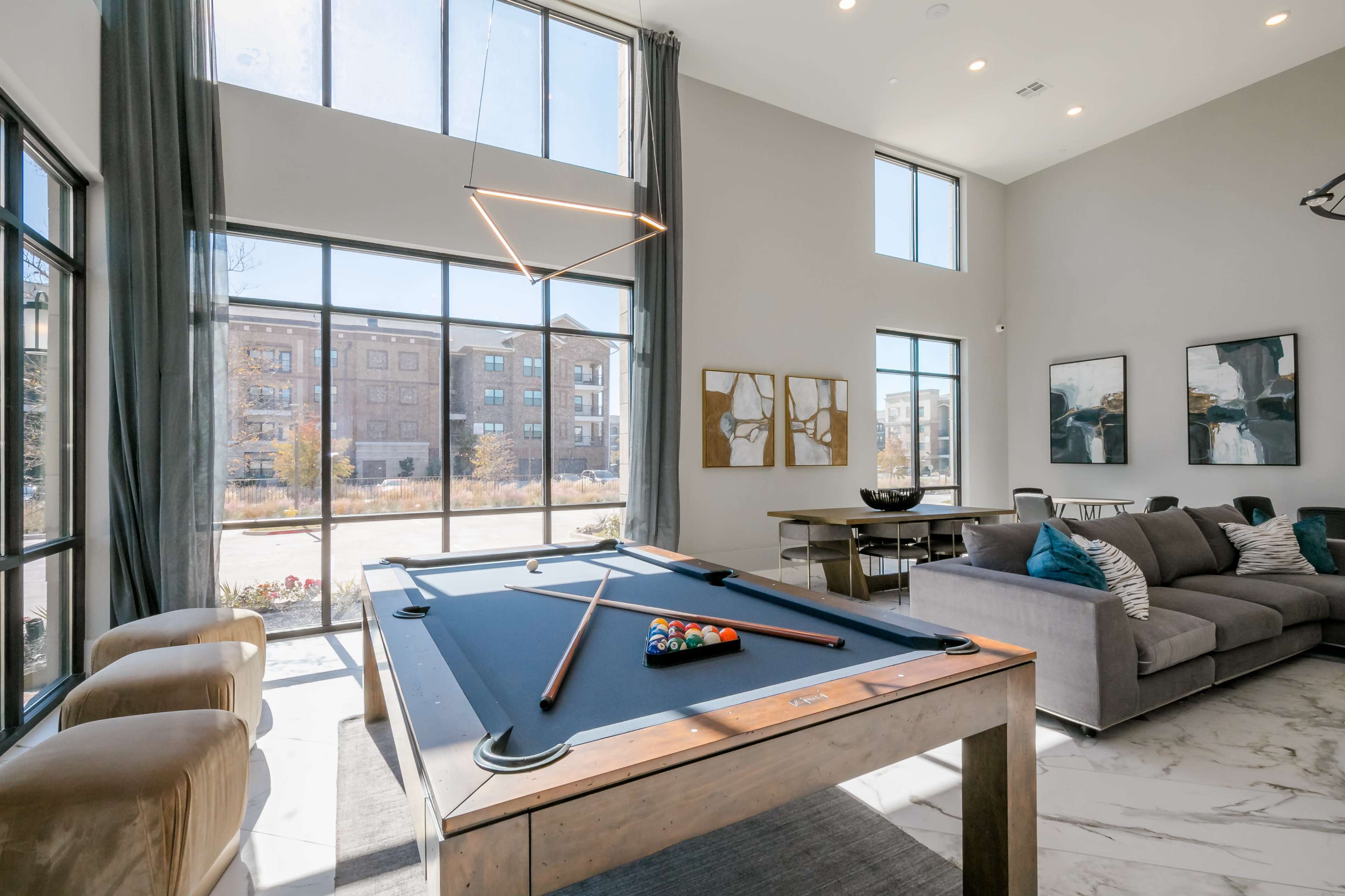 The image shows a modern common living area featuring a pool table, large windows, a sectional couch, and contemporary artwork on the walls.