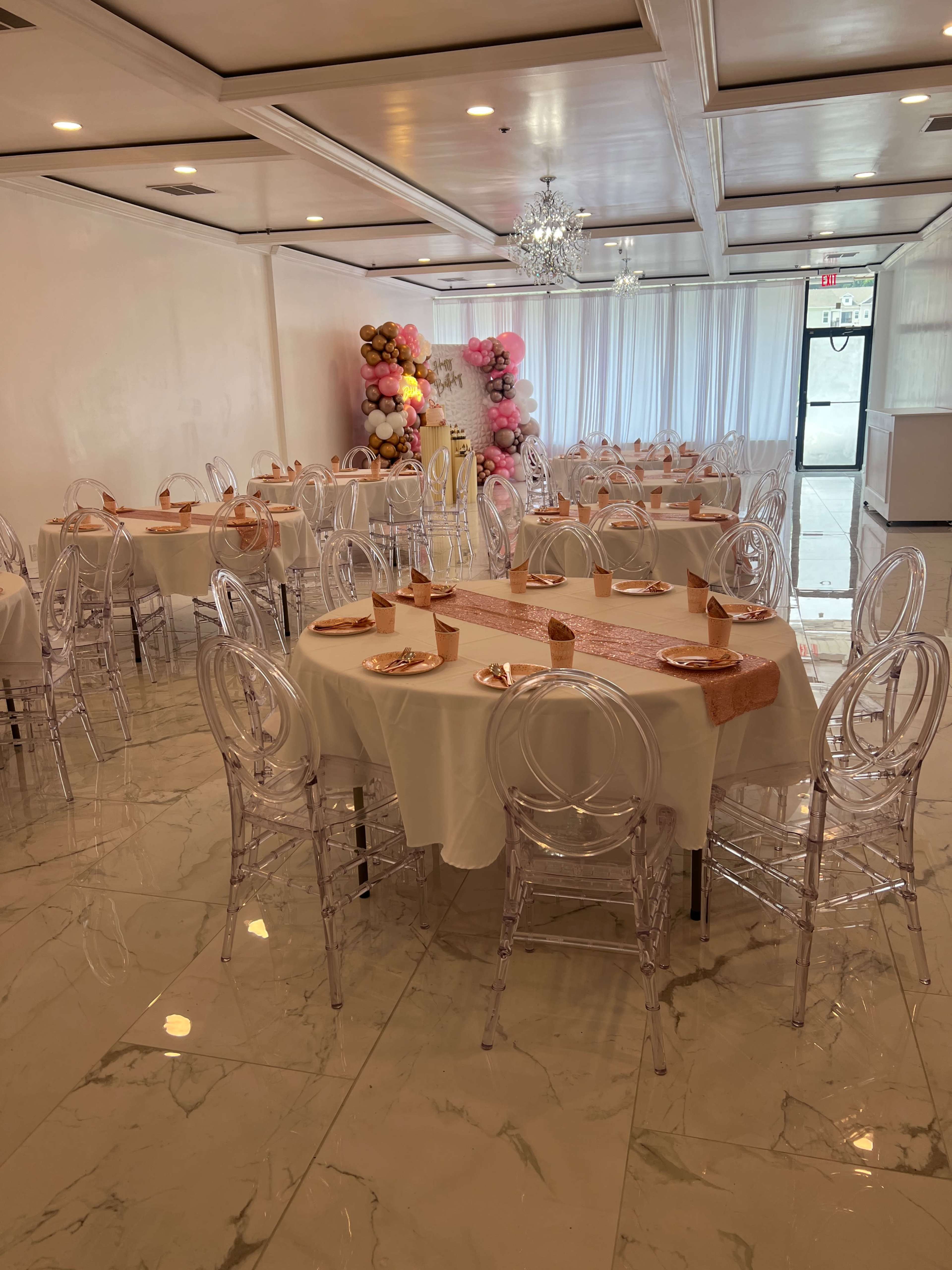 A spacious event room features multiple tables set with white tablecloths and transparent chairs, alongside a pastel balloon display in the corner.