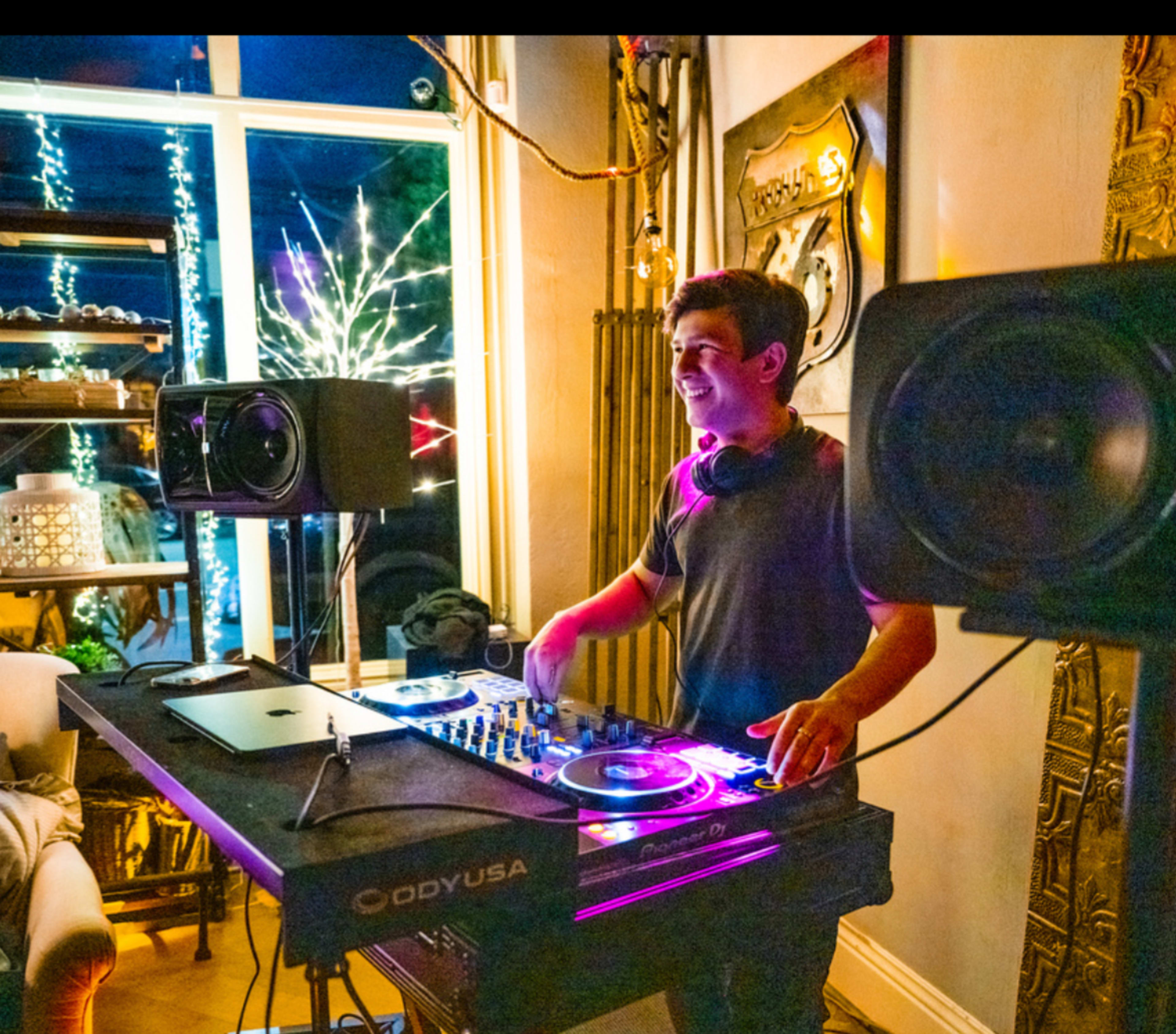 A DJ stands behind a mixing console with turntables and speakers, performing in a softly lit room decorated for an event.