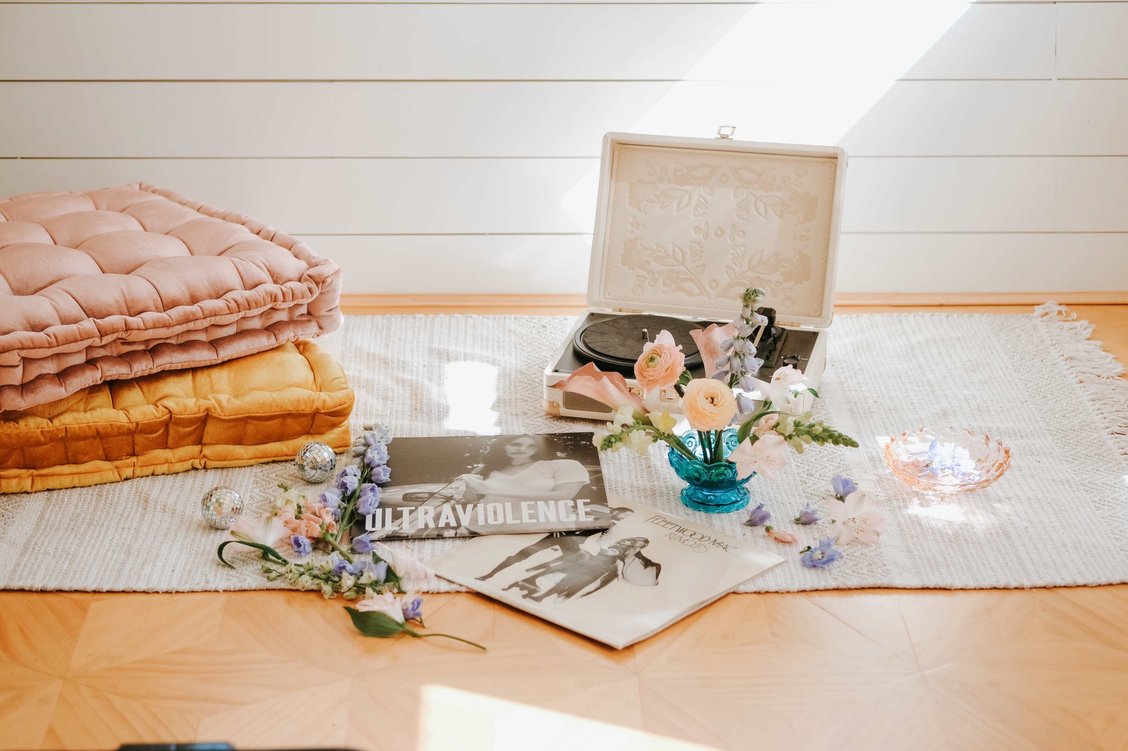 A vintage record player sits among vinyl records, flowers in a vase, and decorative items on a textured rug.