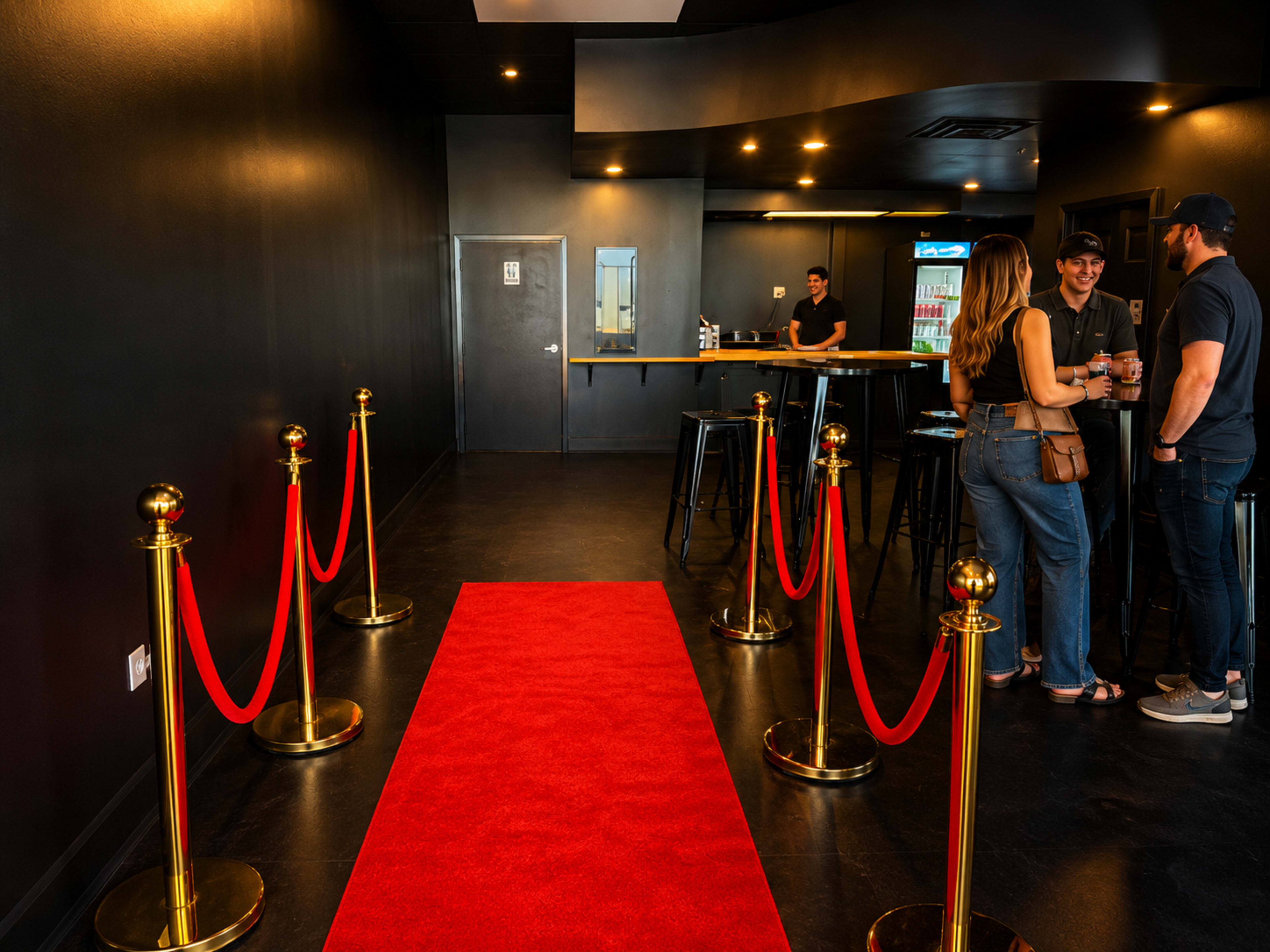 The interior of a venue features a red carpet with velvet ropes, leading to a bar area where three people are engaged in conversation.