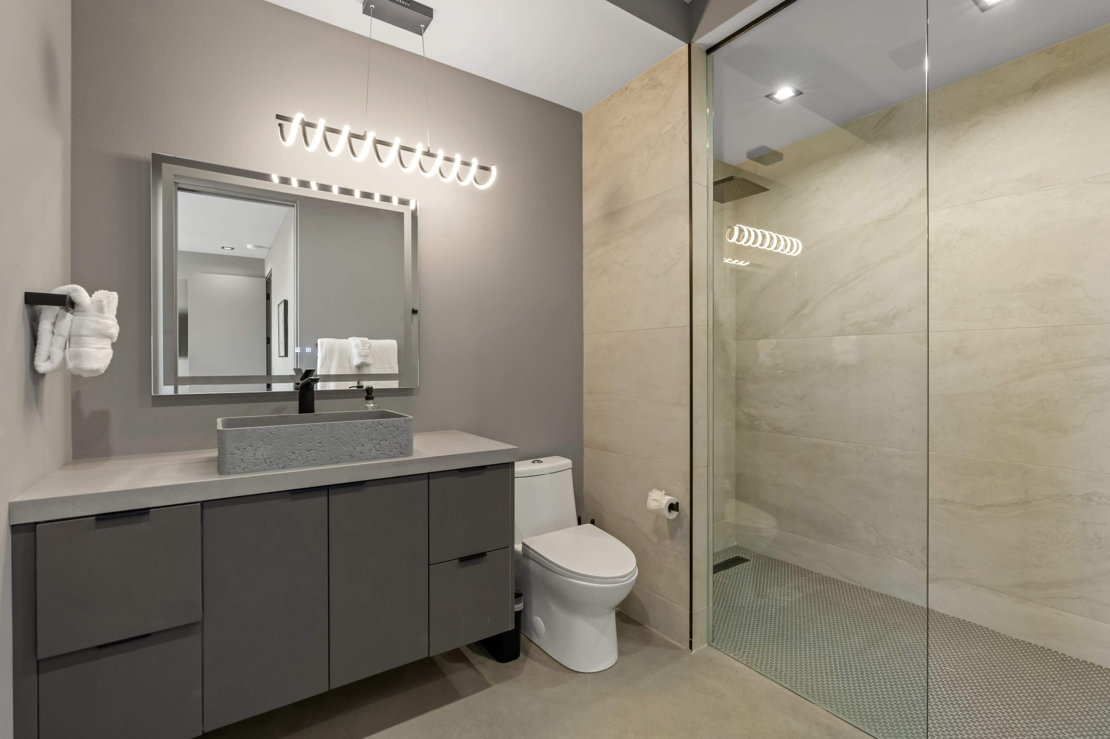 The image shows a modern bathroom featuring a glass shower, a gray vanity with a rectangular sink, and a wall-mounted mirror above the sink.