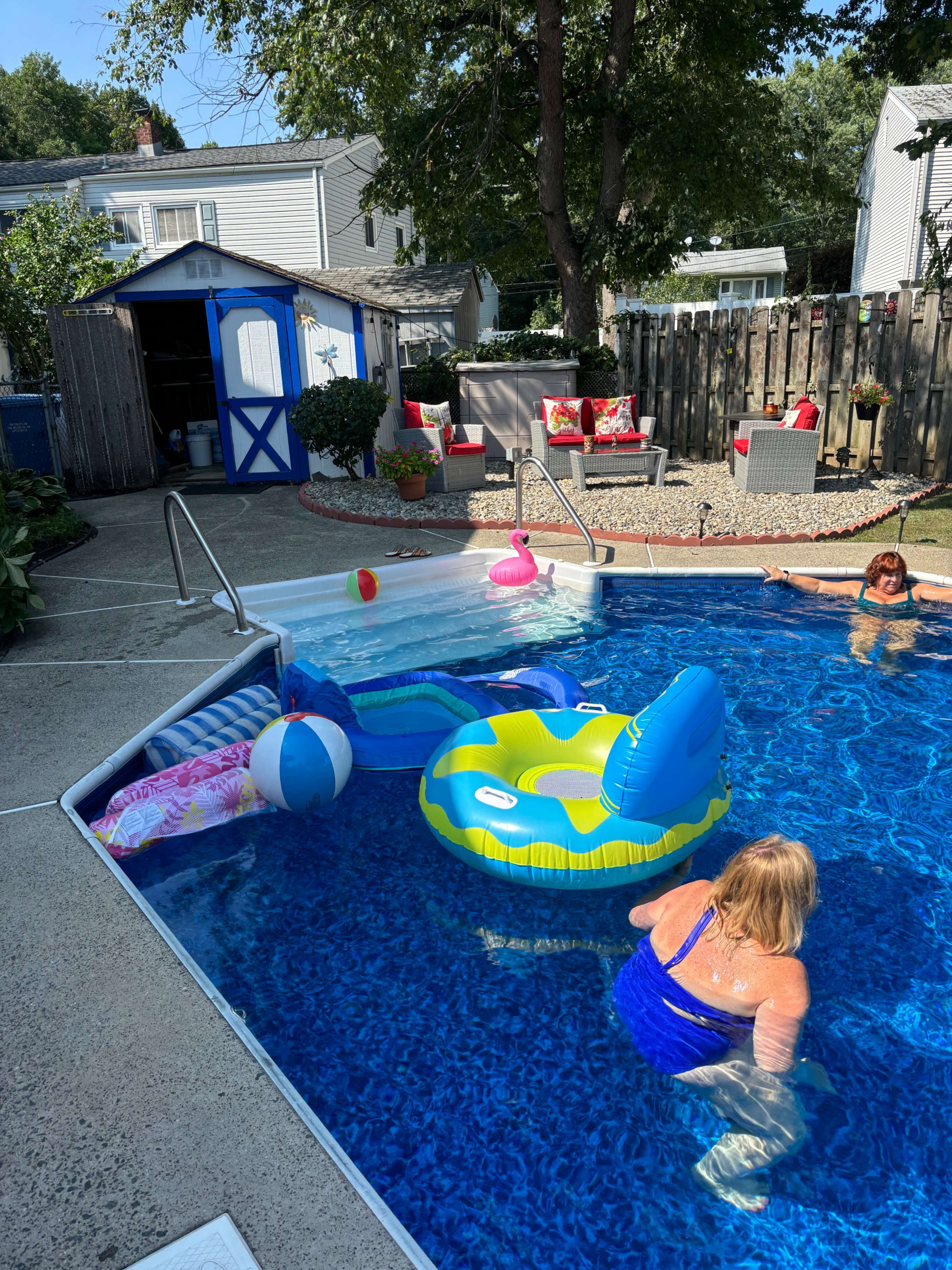 A woman swims in a pool surrounded by inflatable floats, while another person relaxes nearby in a backyard with a shed and seating.
