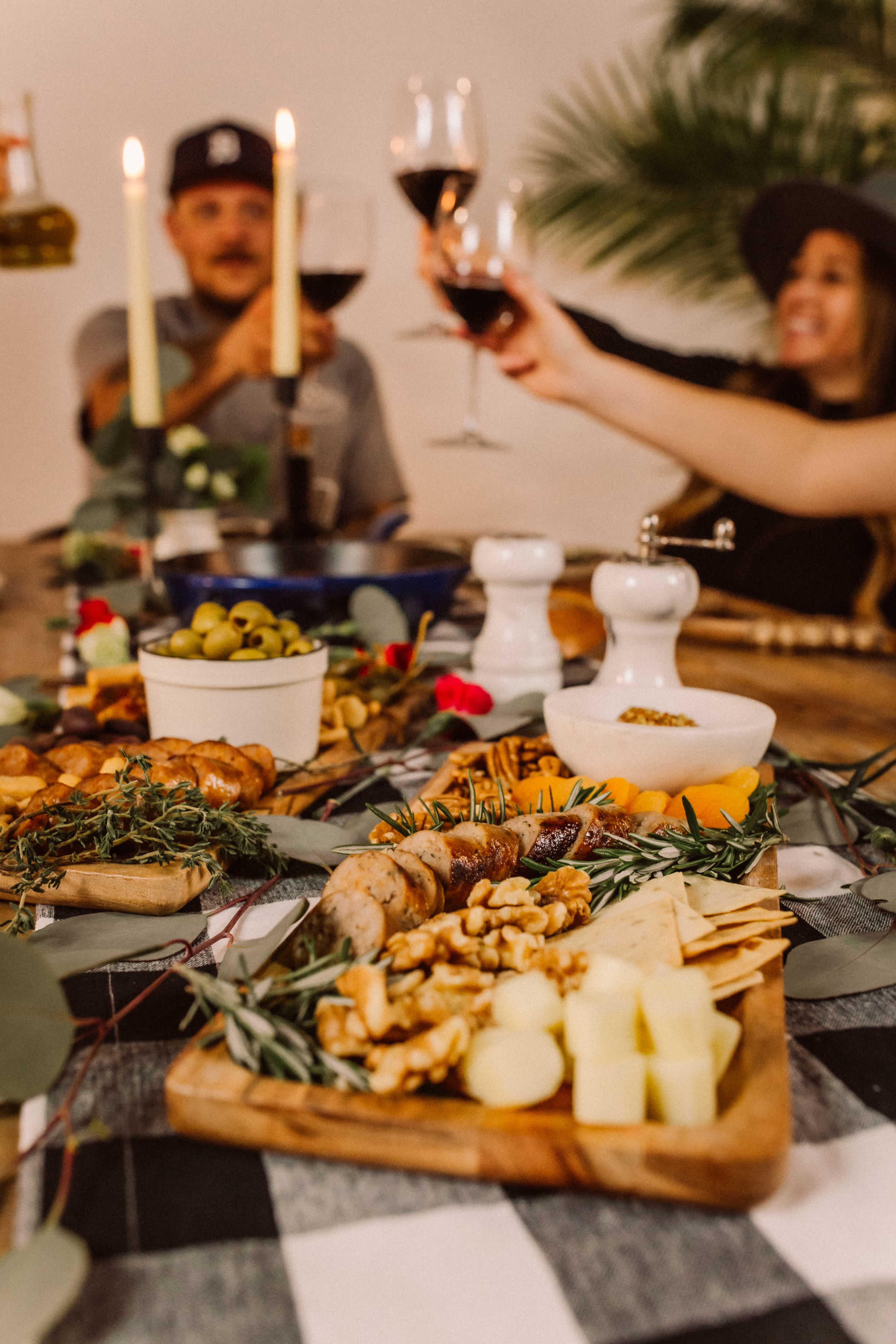 A wooden table is set with a variety of cheeses, nuts, and olives, while two people in the background raise glasses of red wine in a toast.