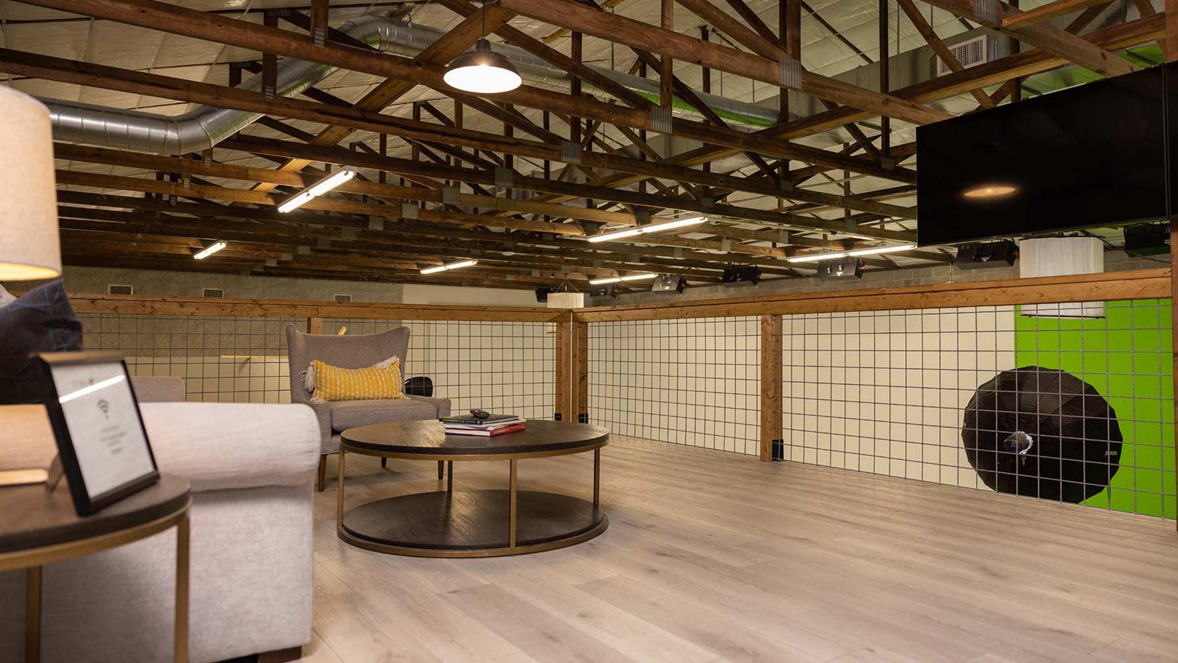 The image shows a lofted space with wooden beams, a circular table and chairs, and a large television mounted on the wall.