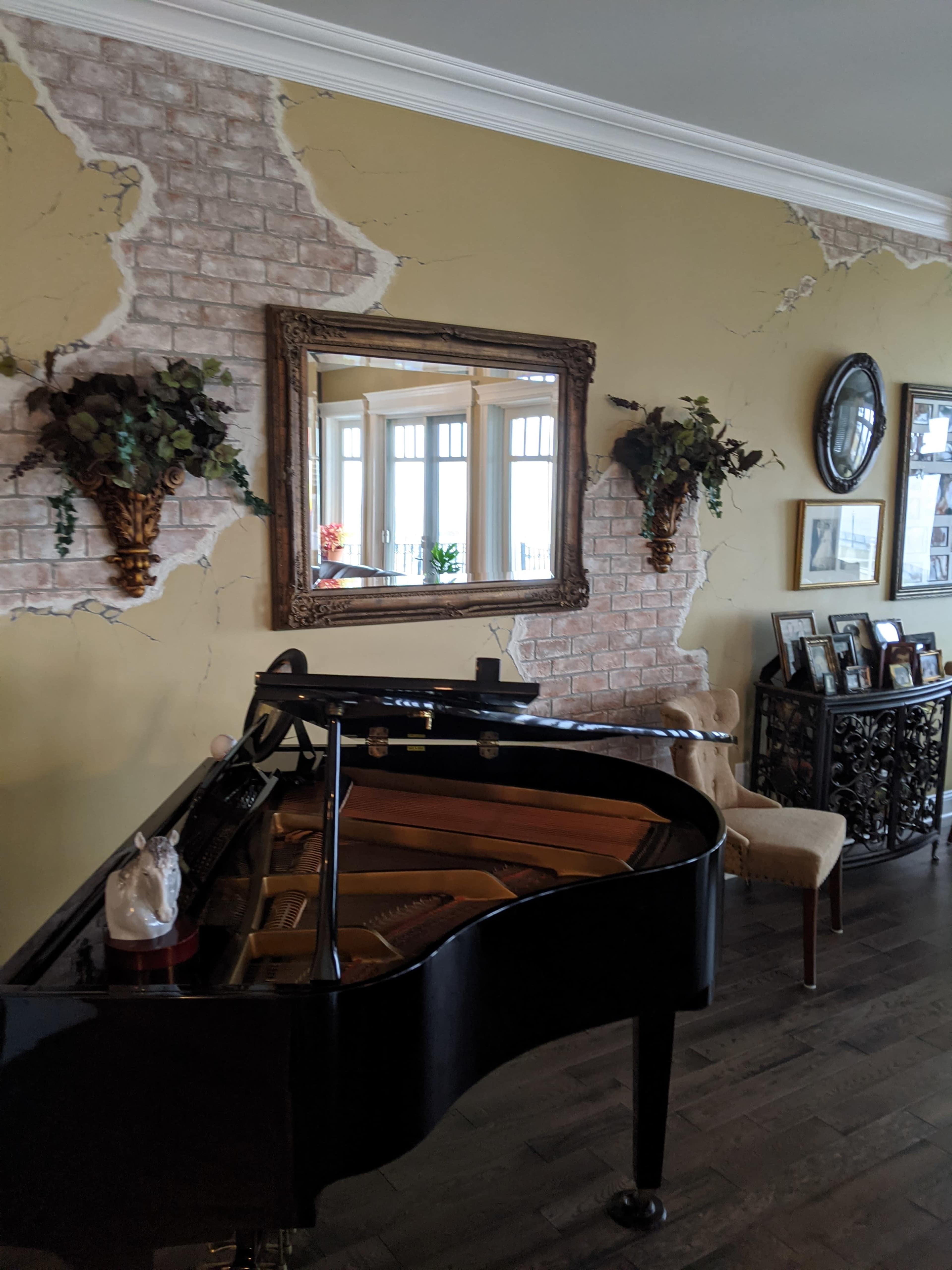 A grand piano sits in front of a wall featuring cracked plaster and decorative elements, including mirrors and wall plants.