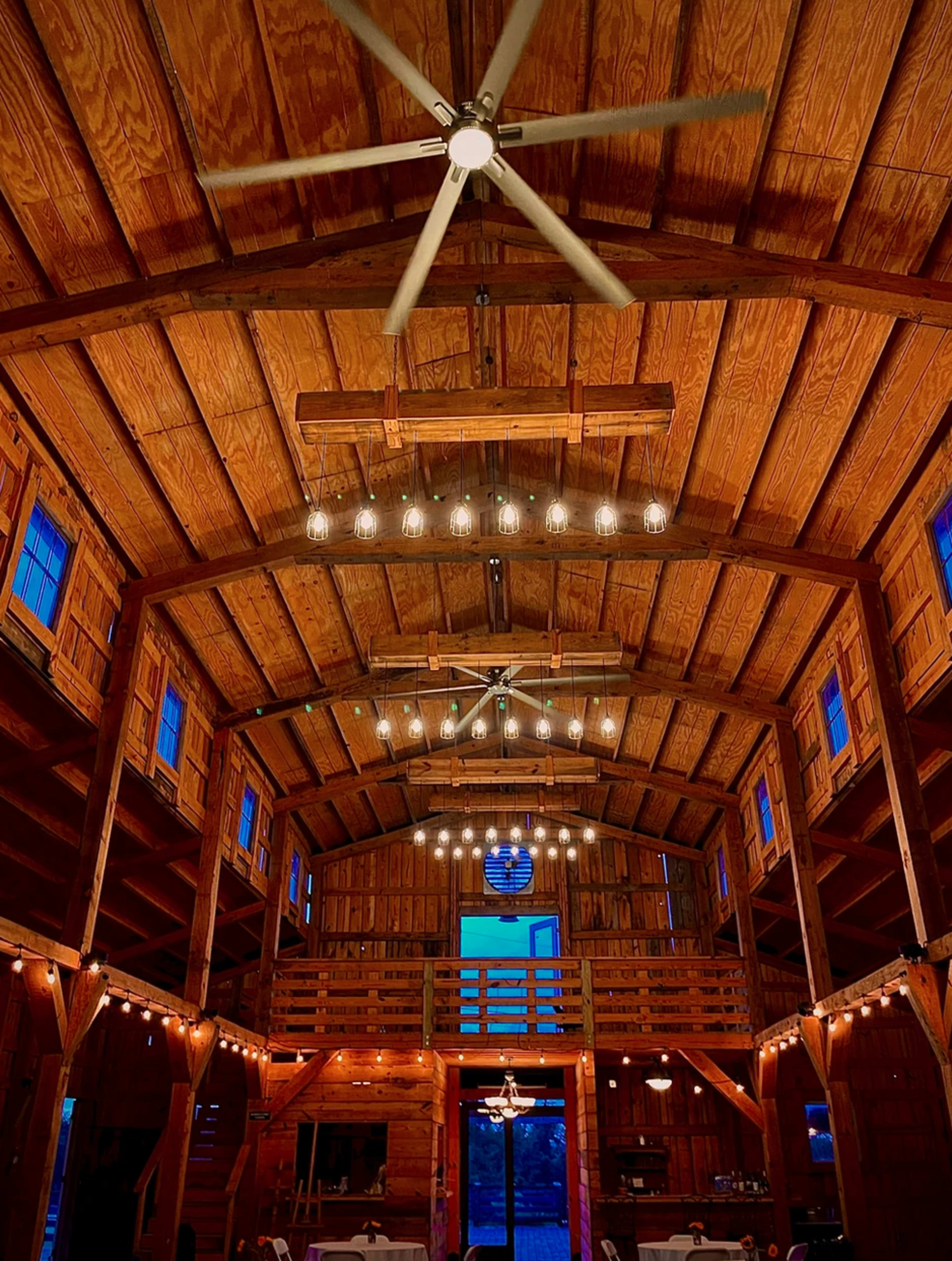 The interior of a large wooden barn features high ceilings, multiple ceiling fans, and string lights illuminating the space.
