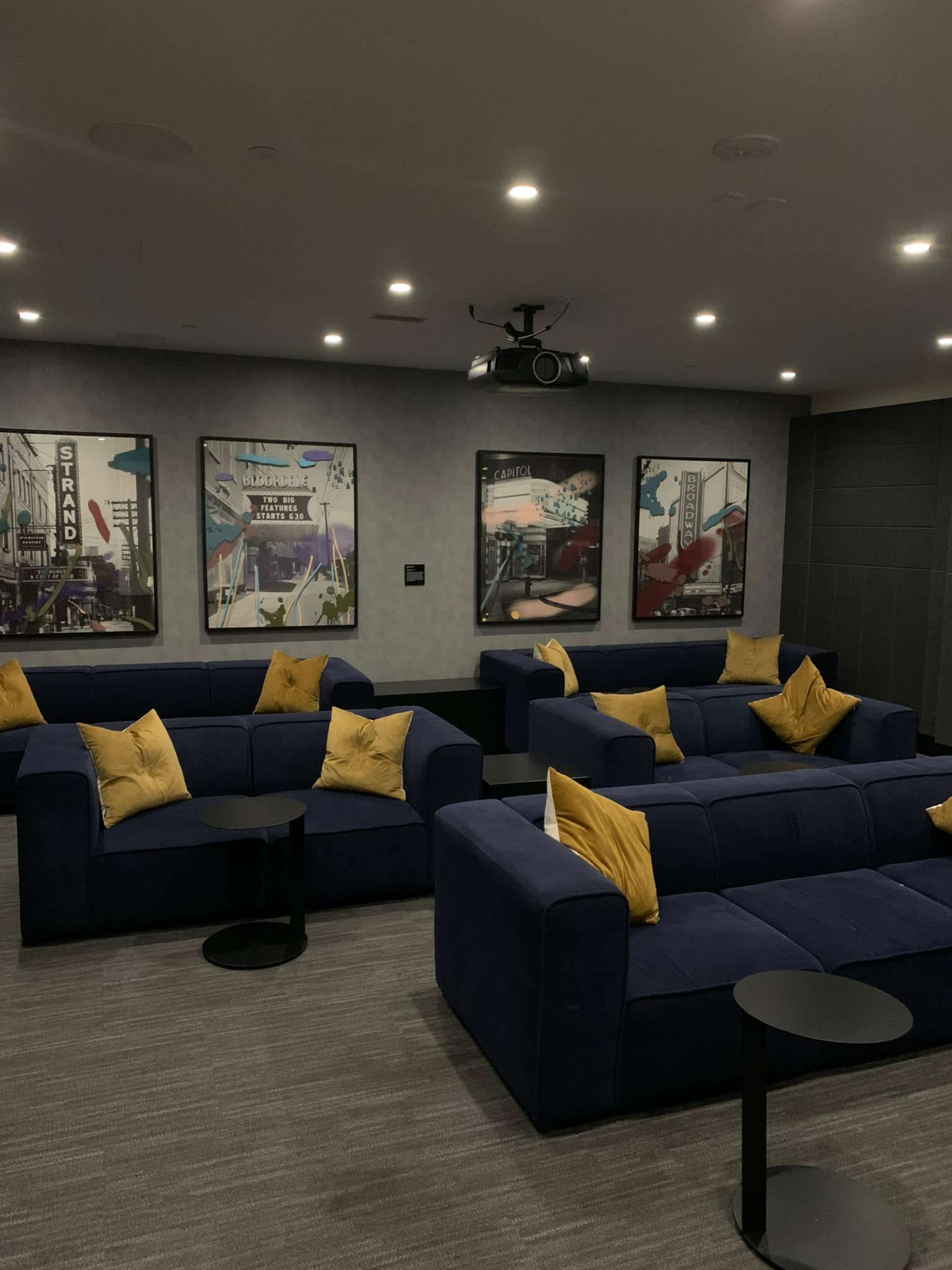 A modern lounge area features blue sofas with yellow cushions arranged around small black tables, complemented by framed art on the walls and a ceiling-mounted projector.