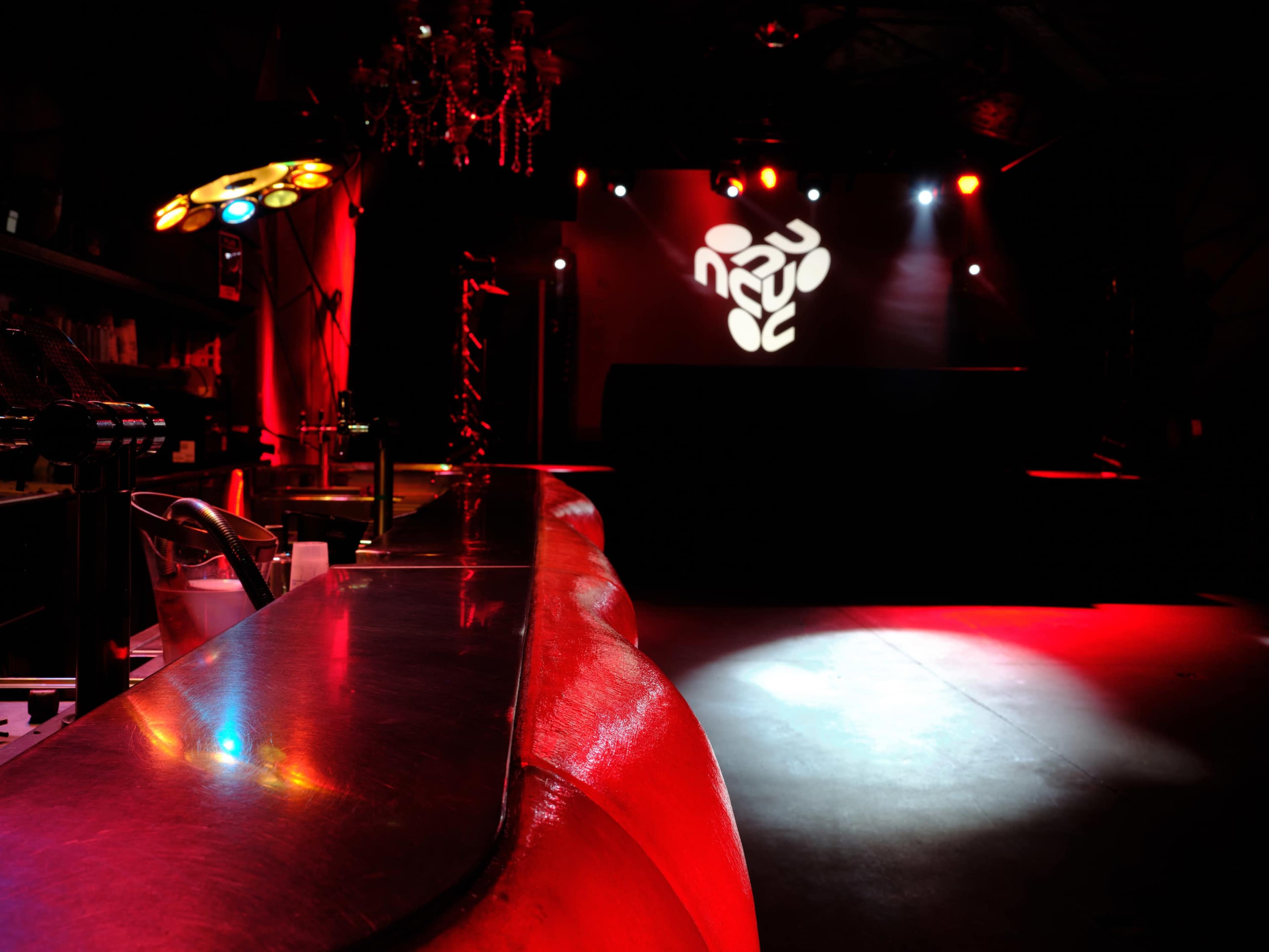 The scene depicts an empty nightclub interior with a polished bar counter and red lighting illuminating the stage area.