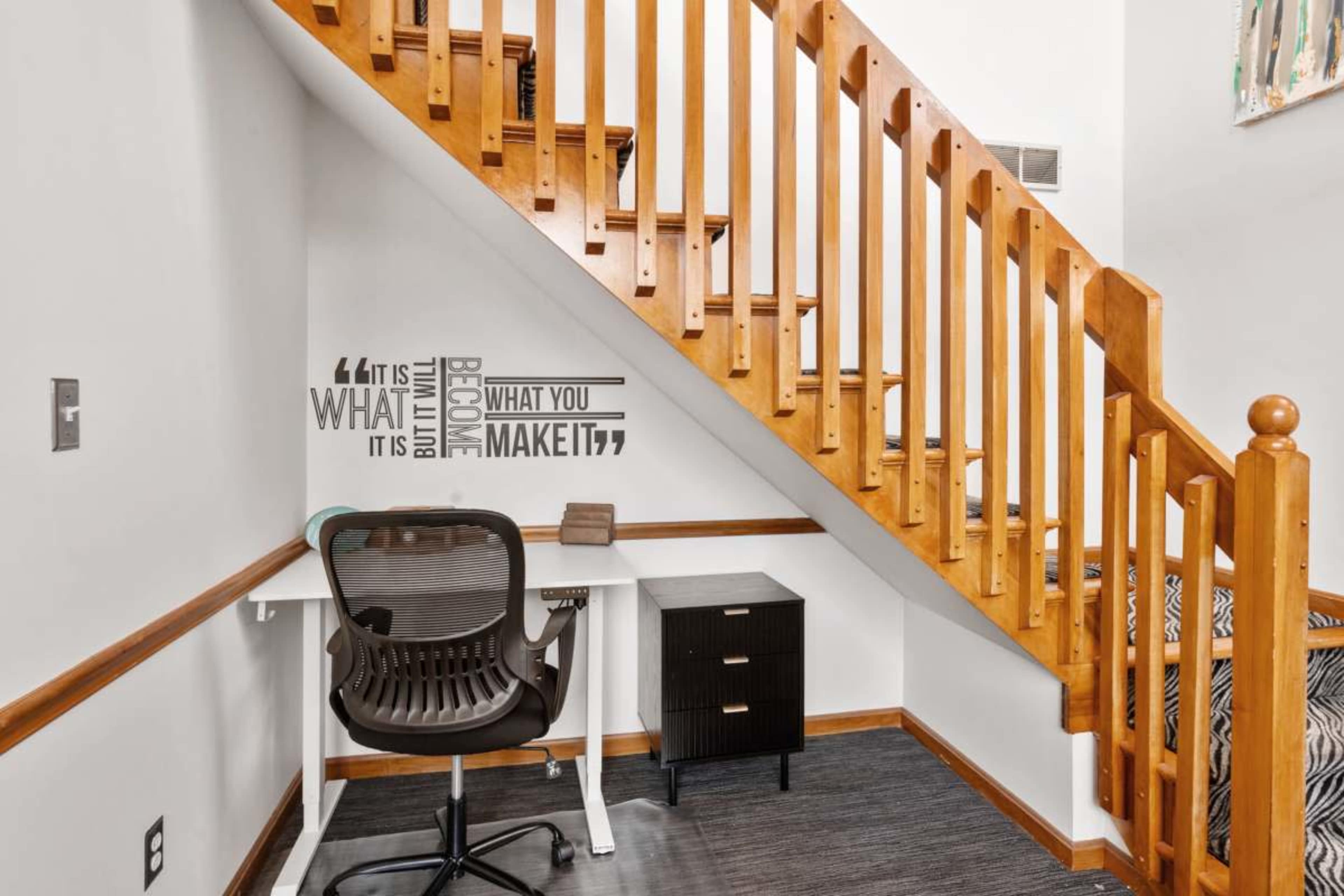 The image shows a small workspace located beneath a staircase, featuring a desk, an ergonomic chair, and a black cabinet, with motivational quotes displayed on the wall.