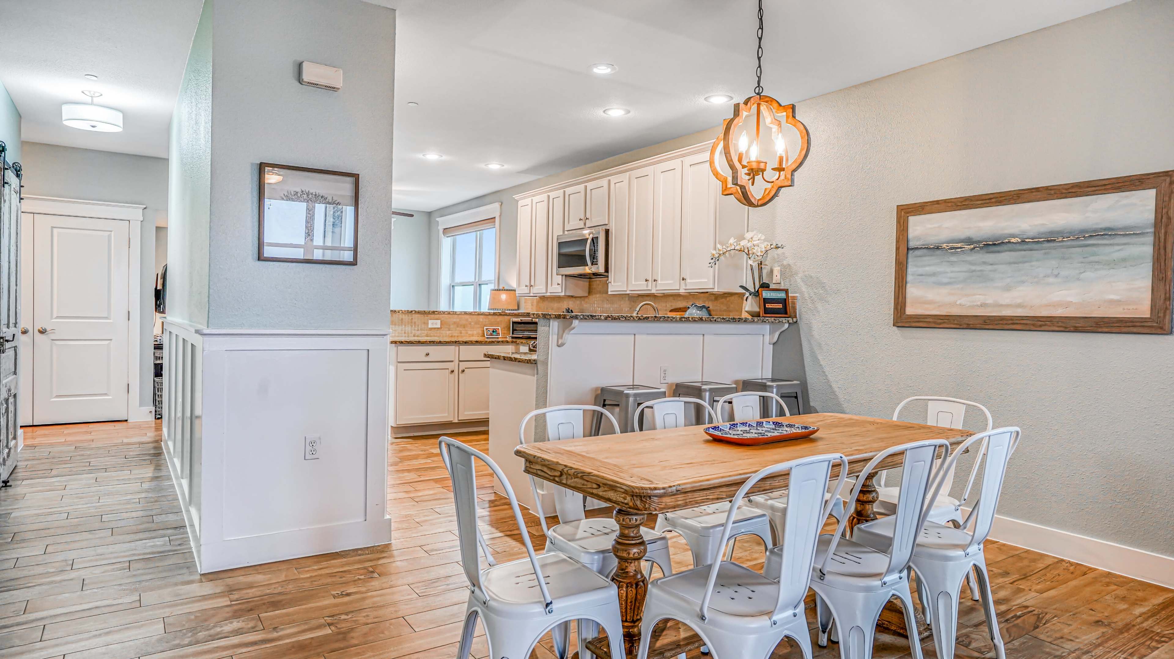 A modern kitchen and dining area with a wooden table and white metal chairs, featuring light-colored cabinetry and a pendant light.