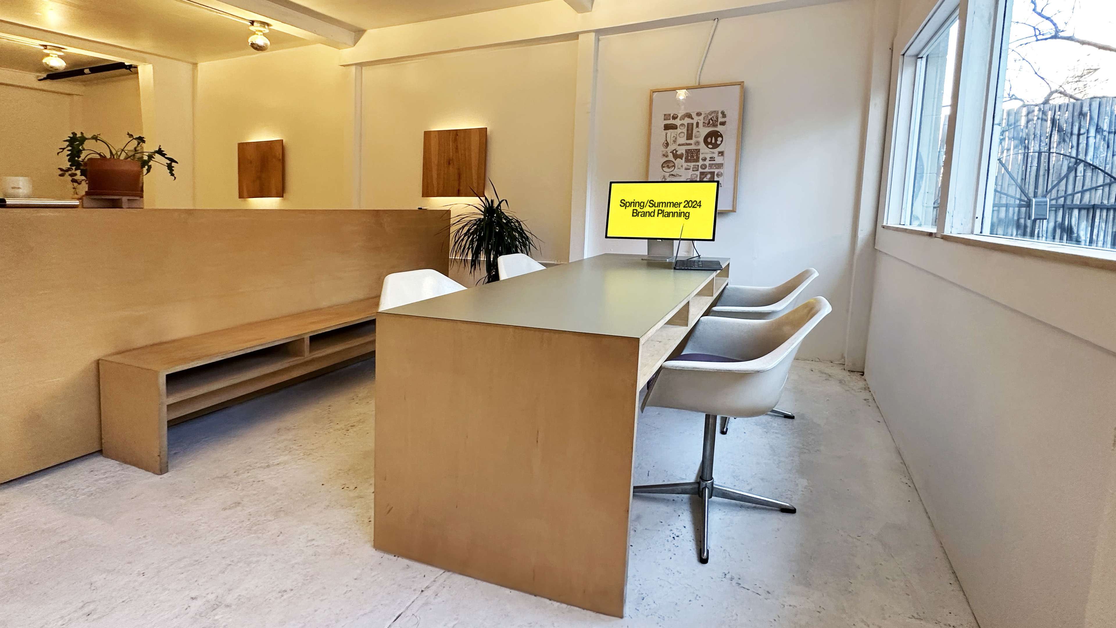 A minimalistic office space with a long wooden table, white chairs, and a monitor displaying "Spring Summer 2024 Brand Planning," surrounded by plants and light-colored walls.