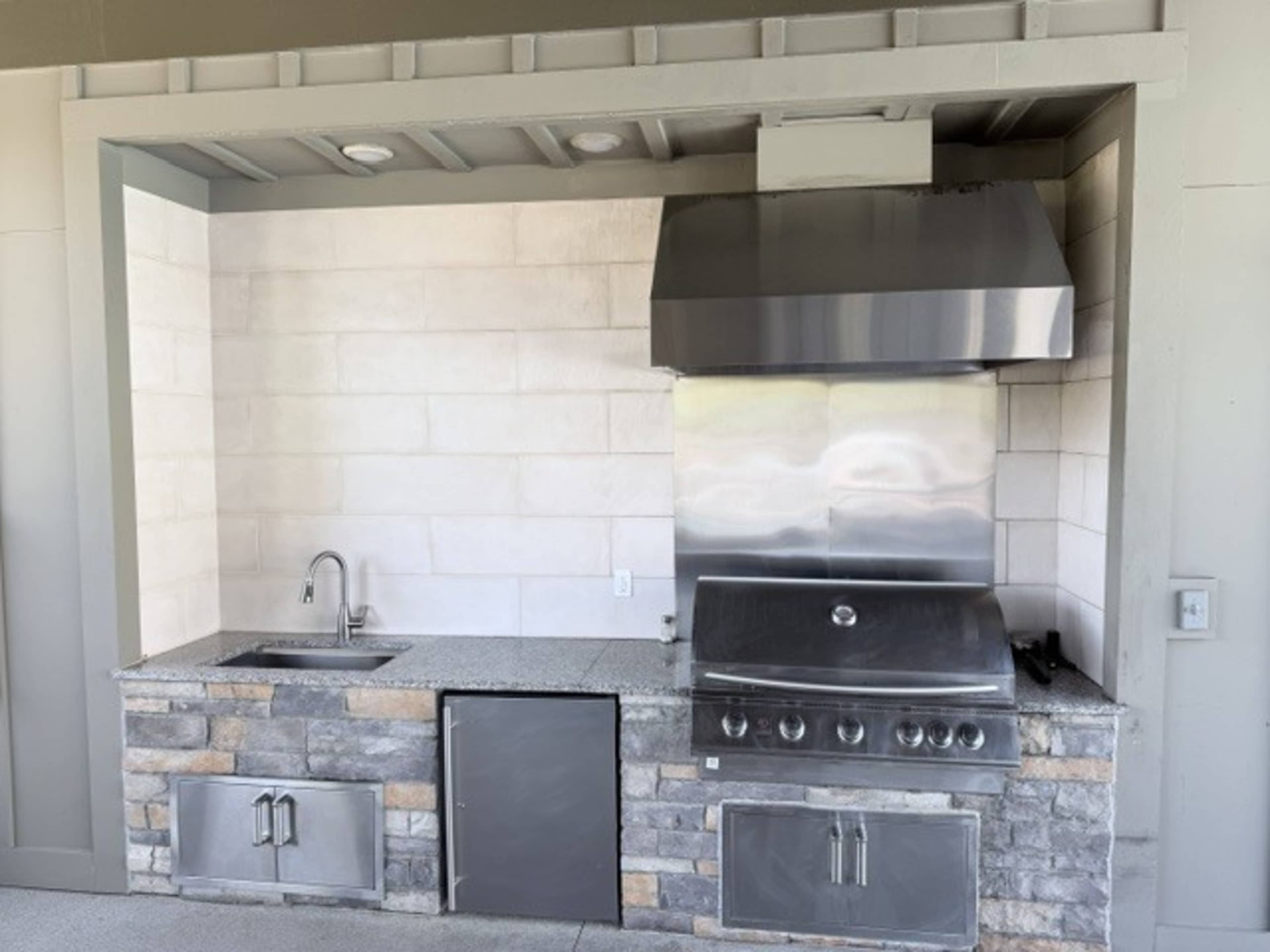 The image shows an outdoor kitchen featuring a stainless steel grill, an exhaust hood, a sink, and a stone counter with a storage cabinet.