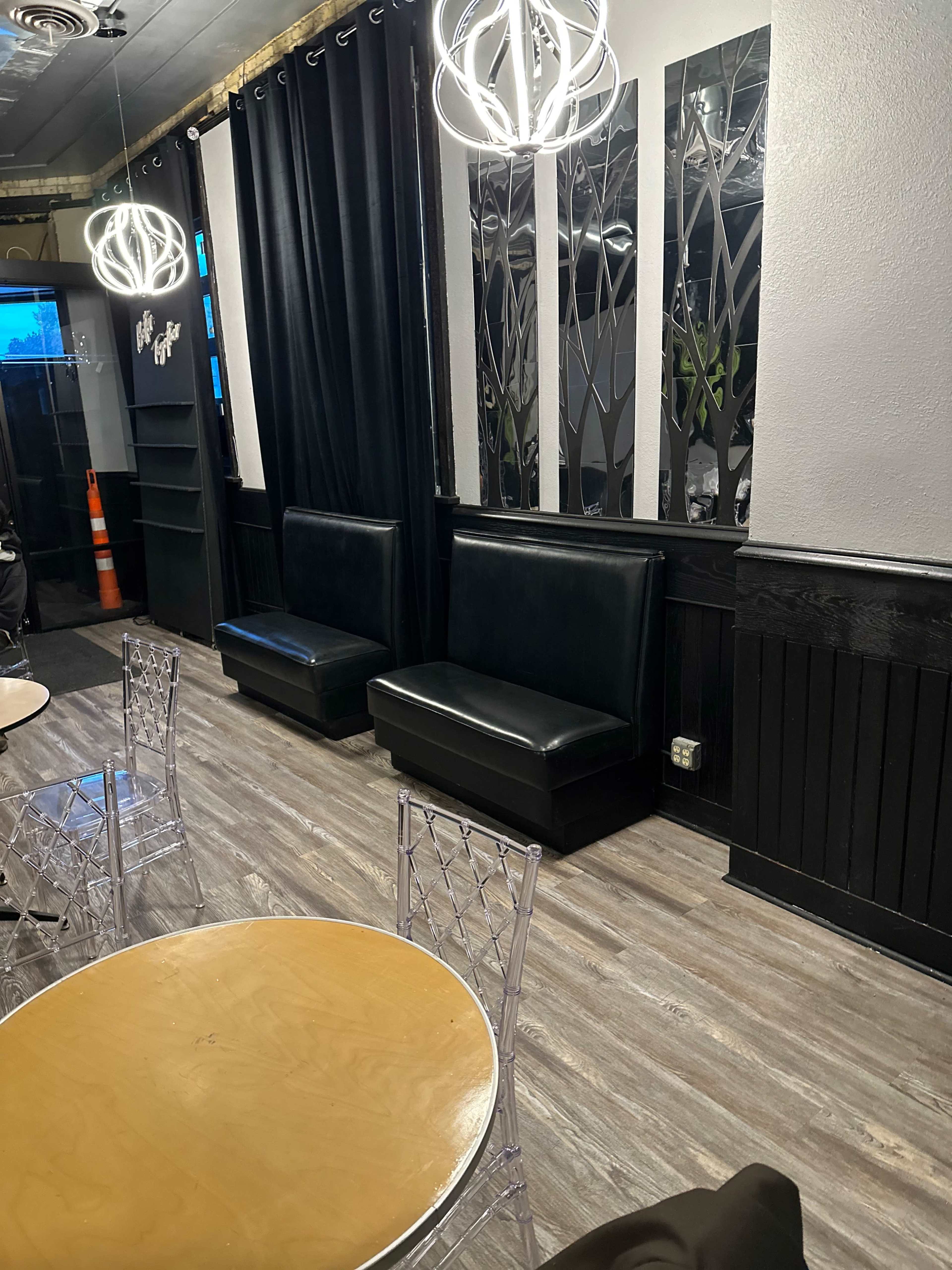 The image shows a modern interior of a café or restaurant featuring two black upholstered benches against a wall, wooden floors, and decorative mirrors.