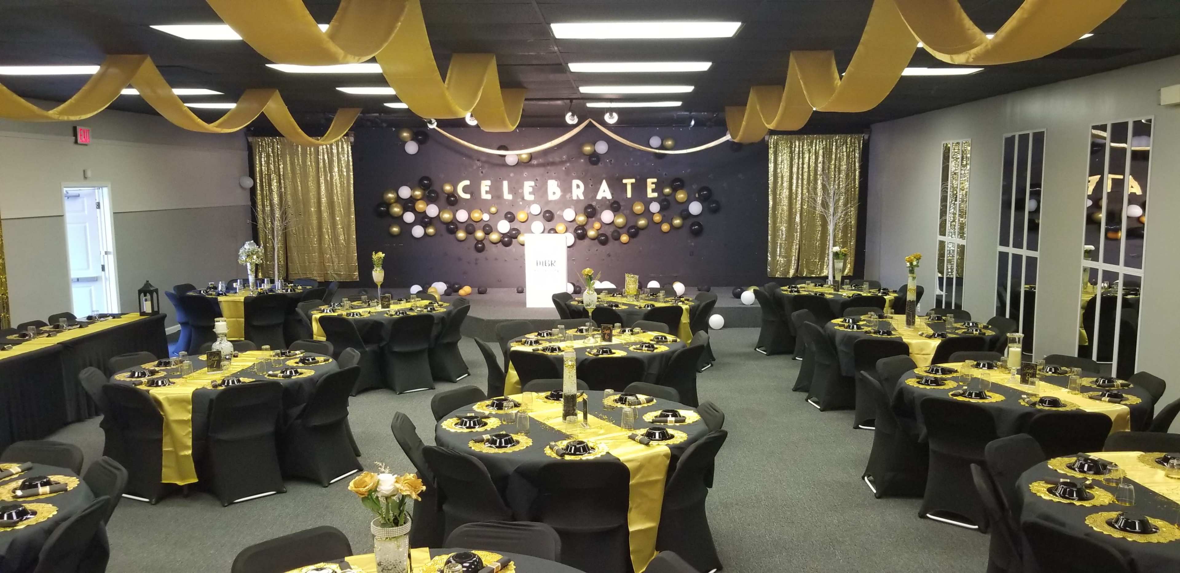 The image shows a decorated event space with black and gold table settings, a backdrop that reads "CELEBRATE," and various balloons and streamers adorning the walls.