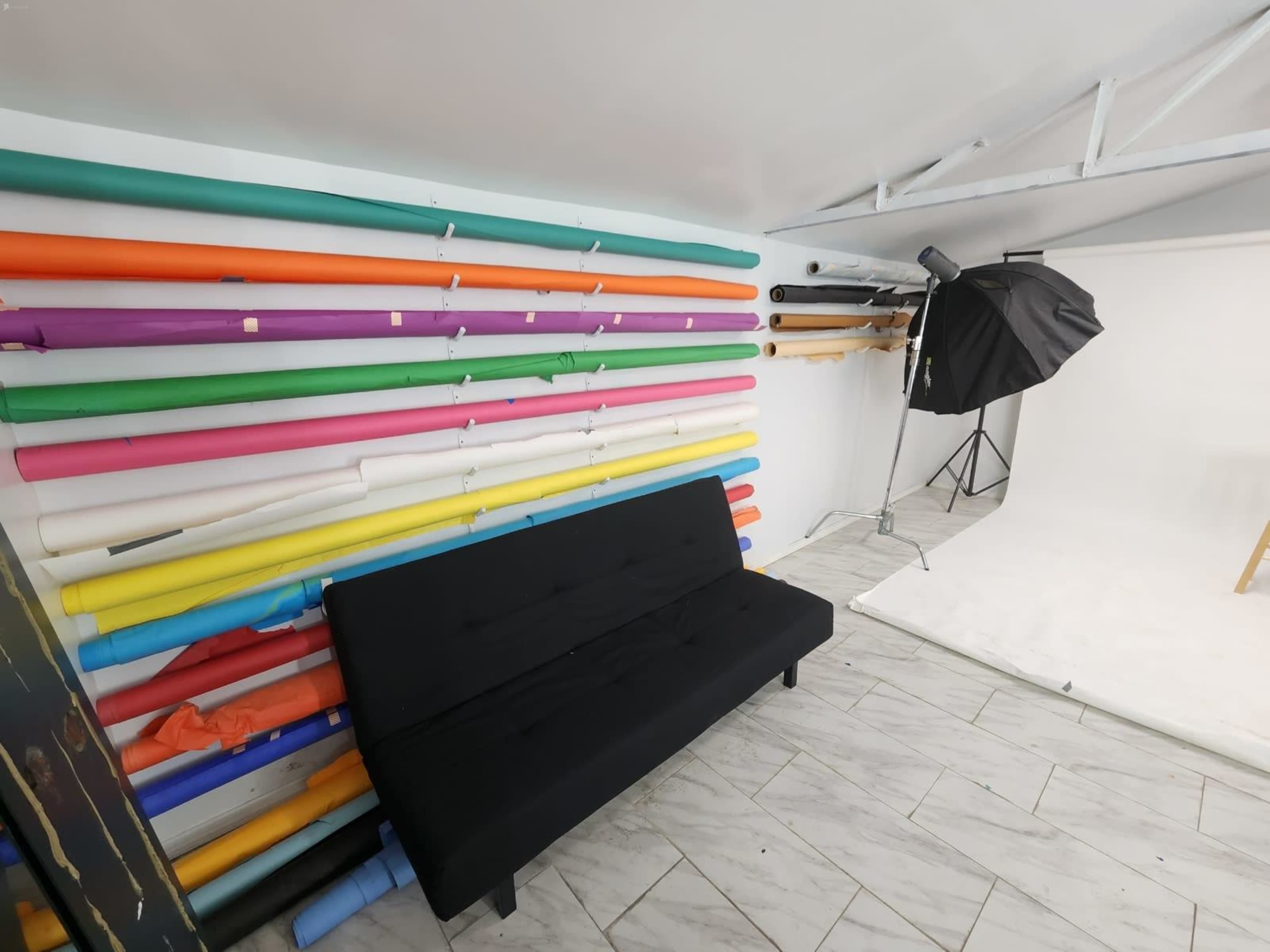 A photography studio with a black couch against a wall lined with colorful rolls of backdrop paper and a lighting setup in the corner.