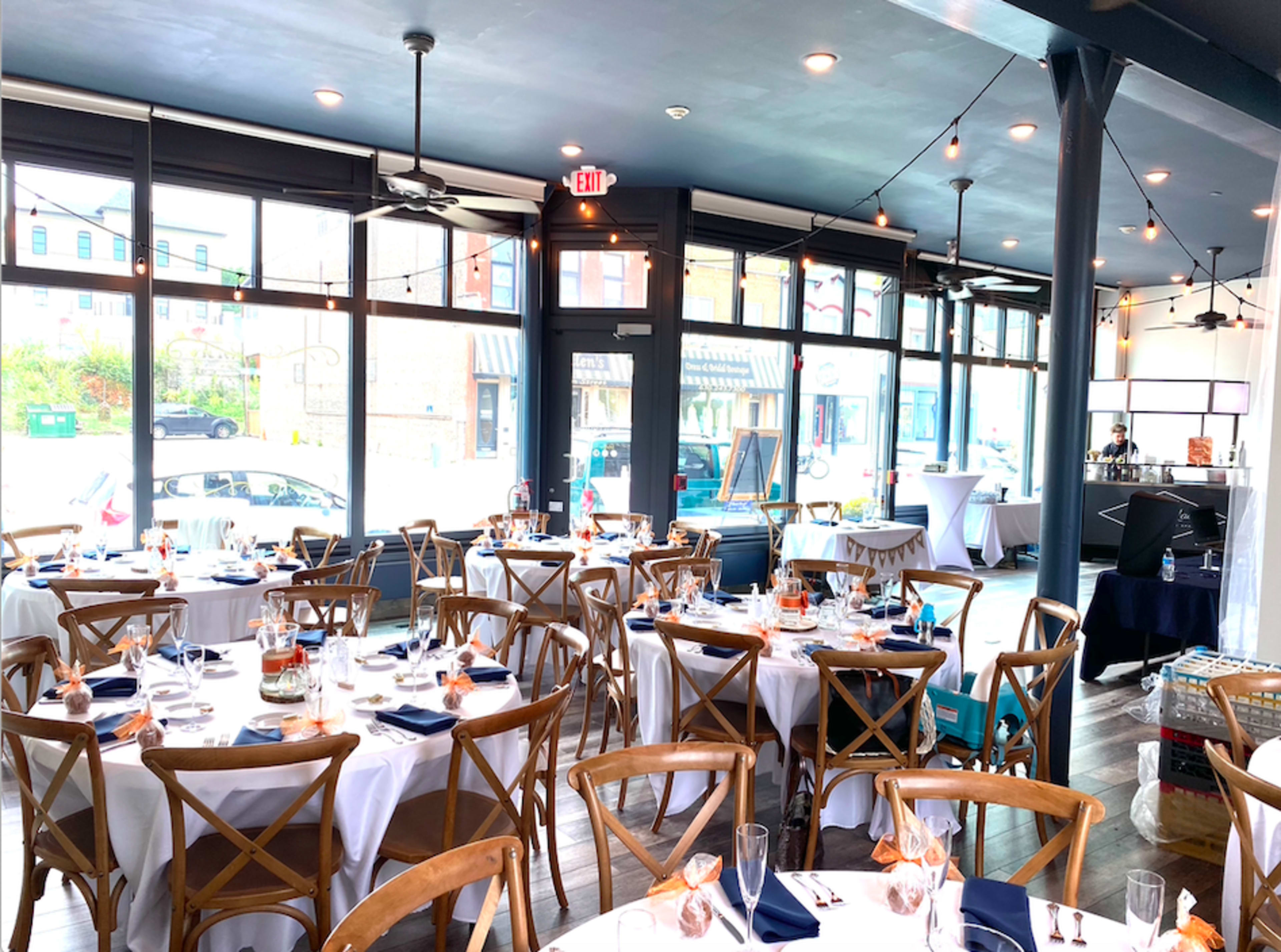 The interior of a restaurant features wooden tables set for dining, with large windows providing natural light and a view of the street outside.