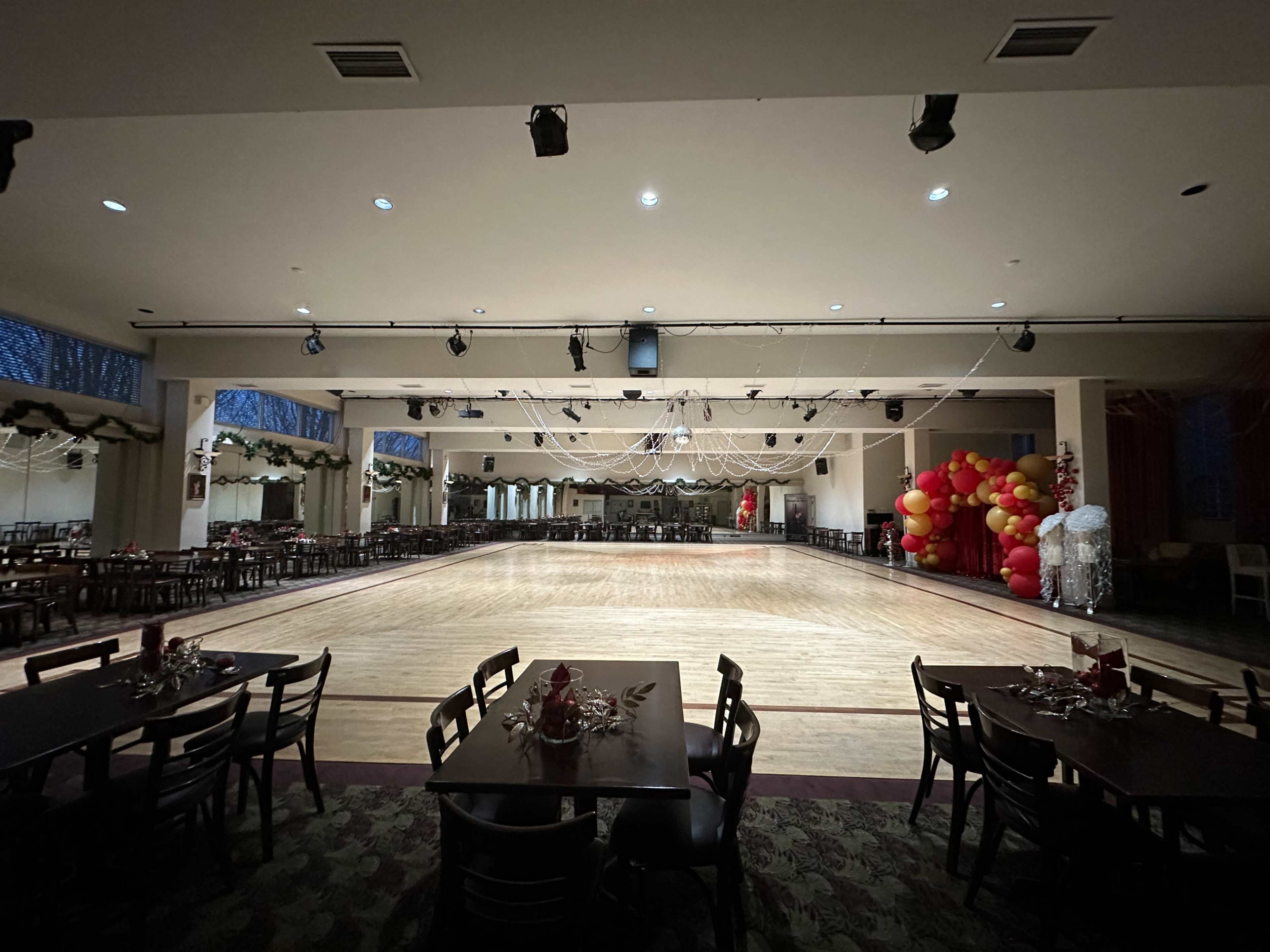 The image shows a spacious event hall with a wooden dance floor, surrounded by tables set for a gathering, and decorated with balloons in one corner.