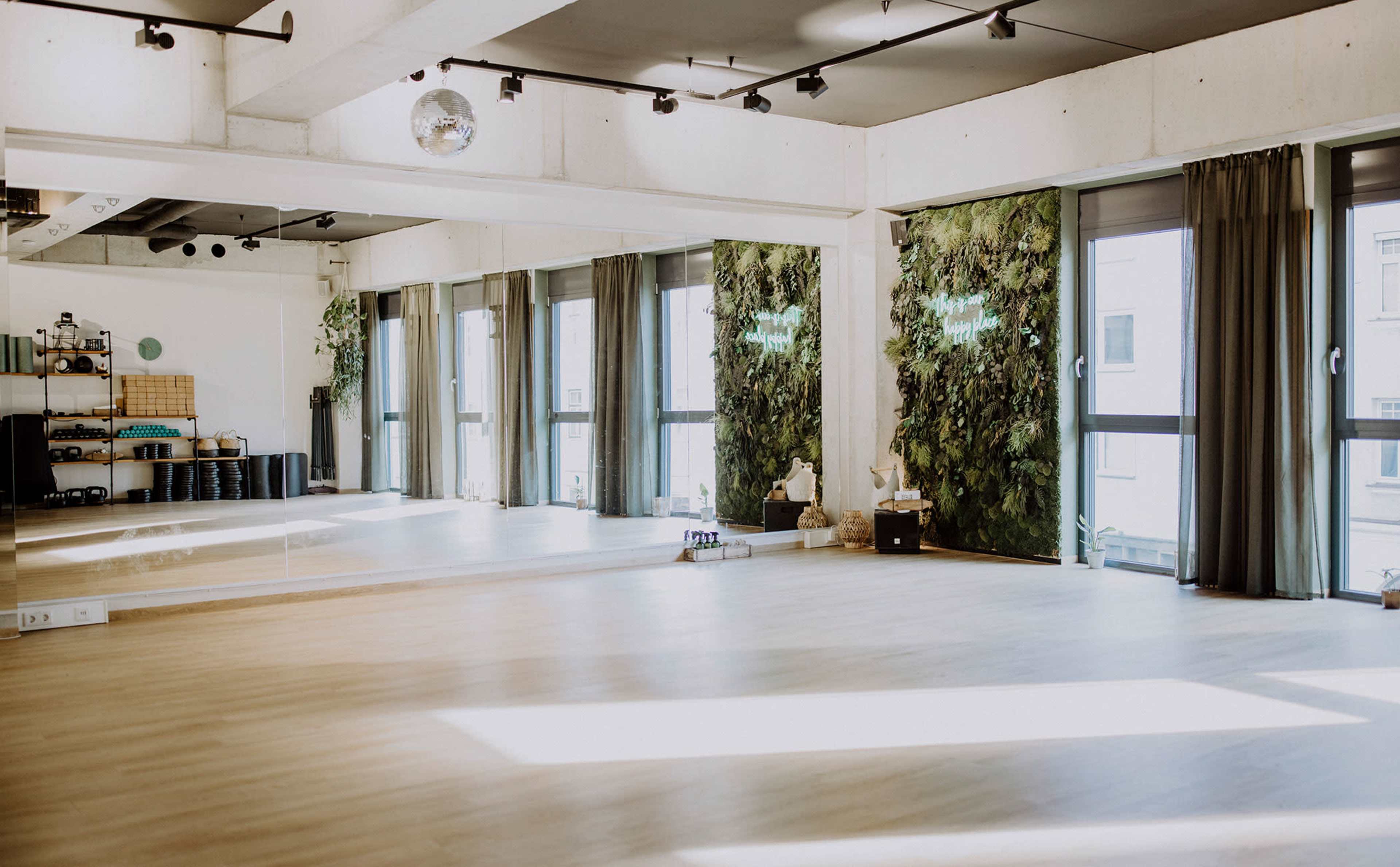 The image shows a spacious studio with large windows, wooden flooring, and green accents along the walls, ideal for activities like yoga or dance.