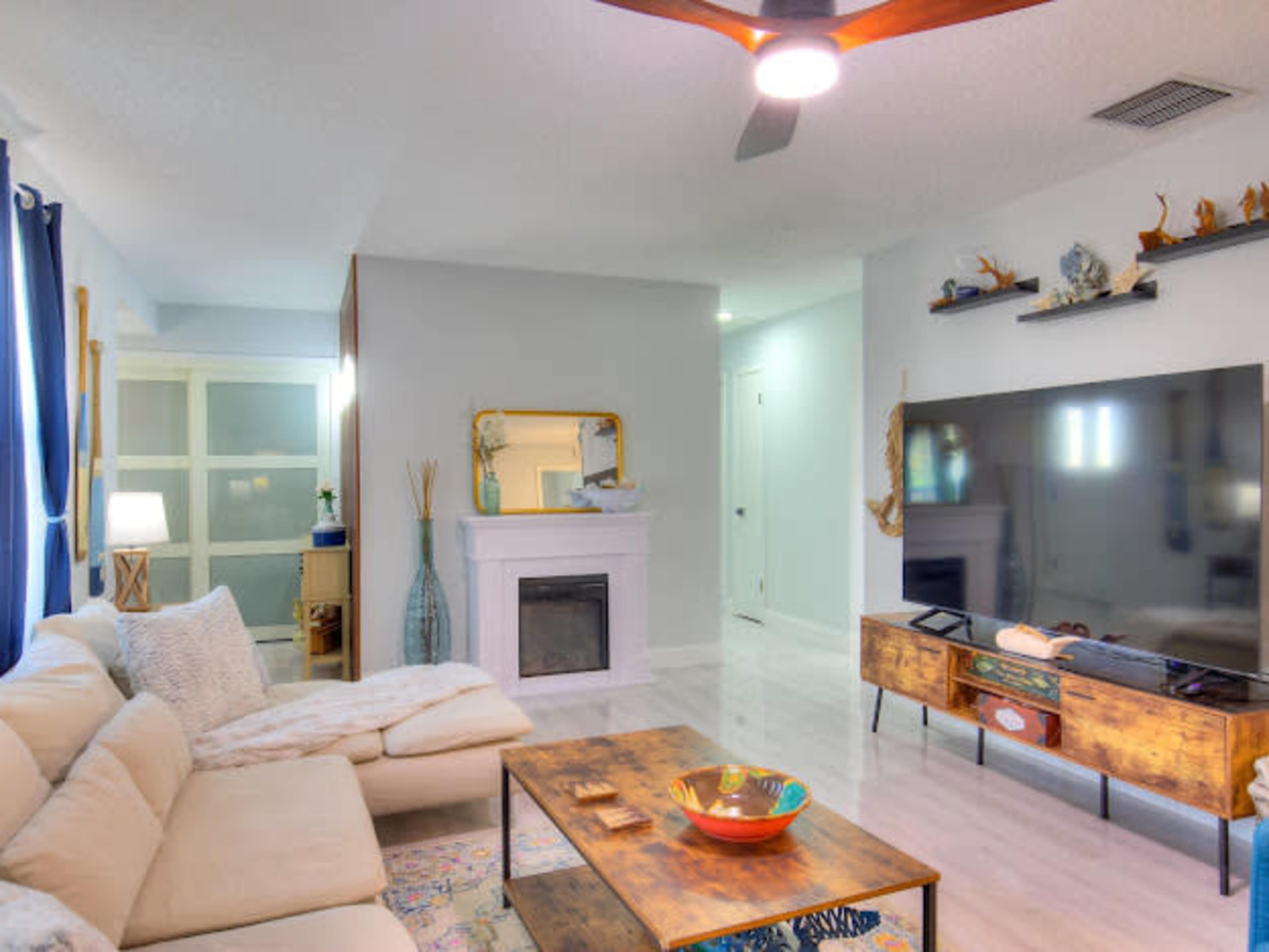 The image shows a modern living room with a beige sectional sofa, a wooden coffee table, a large television on a stand, and a fireplace with a mirror above it.