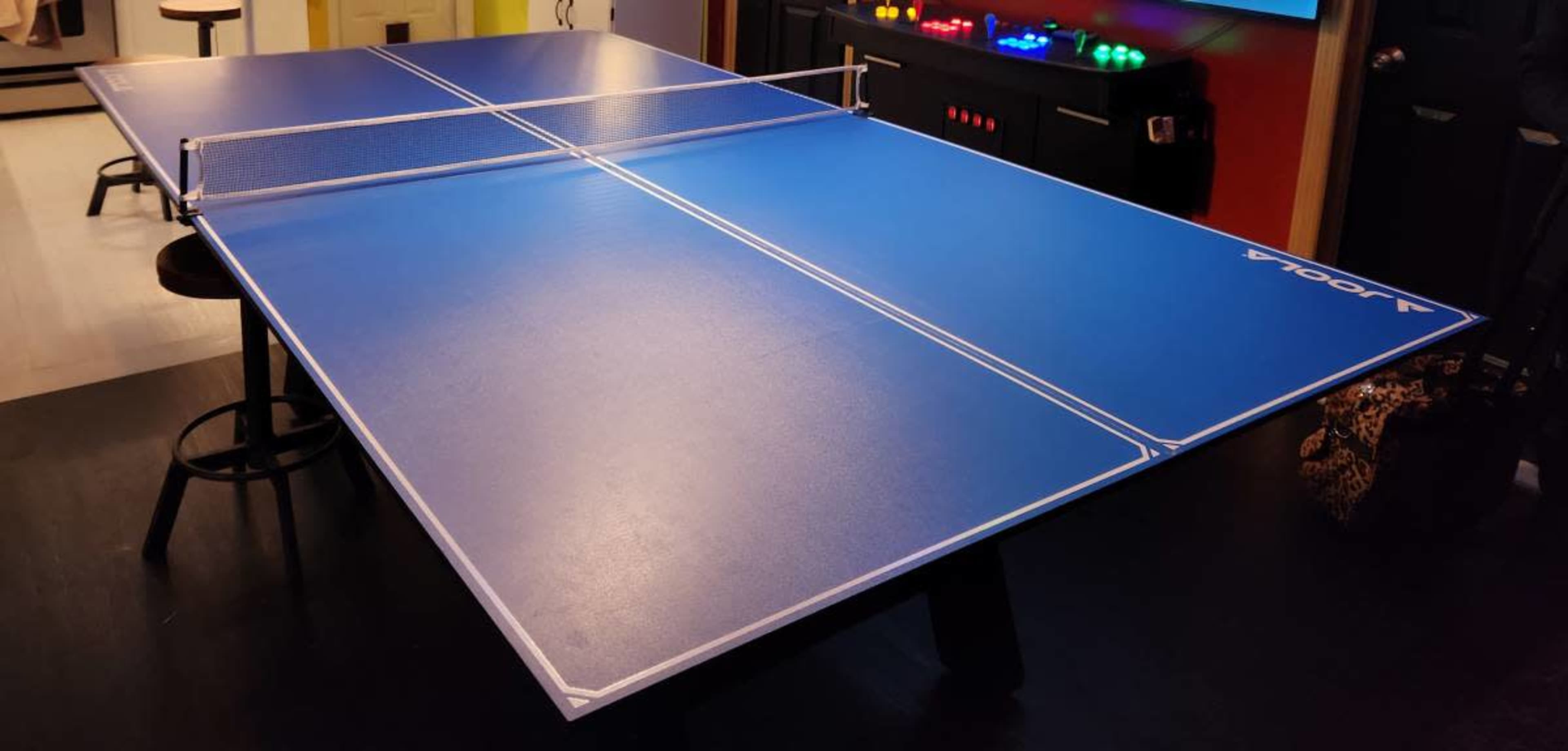 The image shows a blue ping pong table with a net in the center, set in a room with various colored lights in the background.