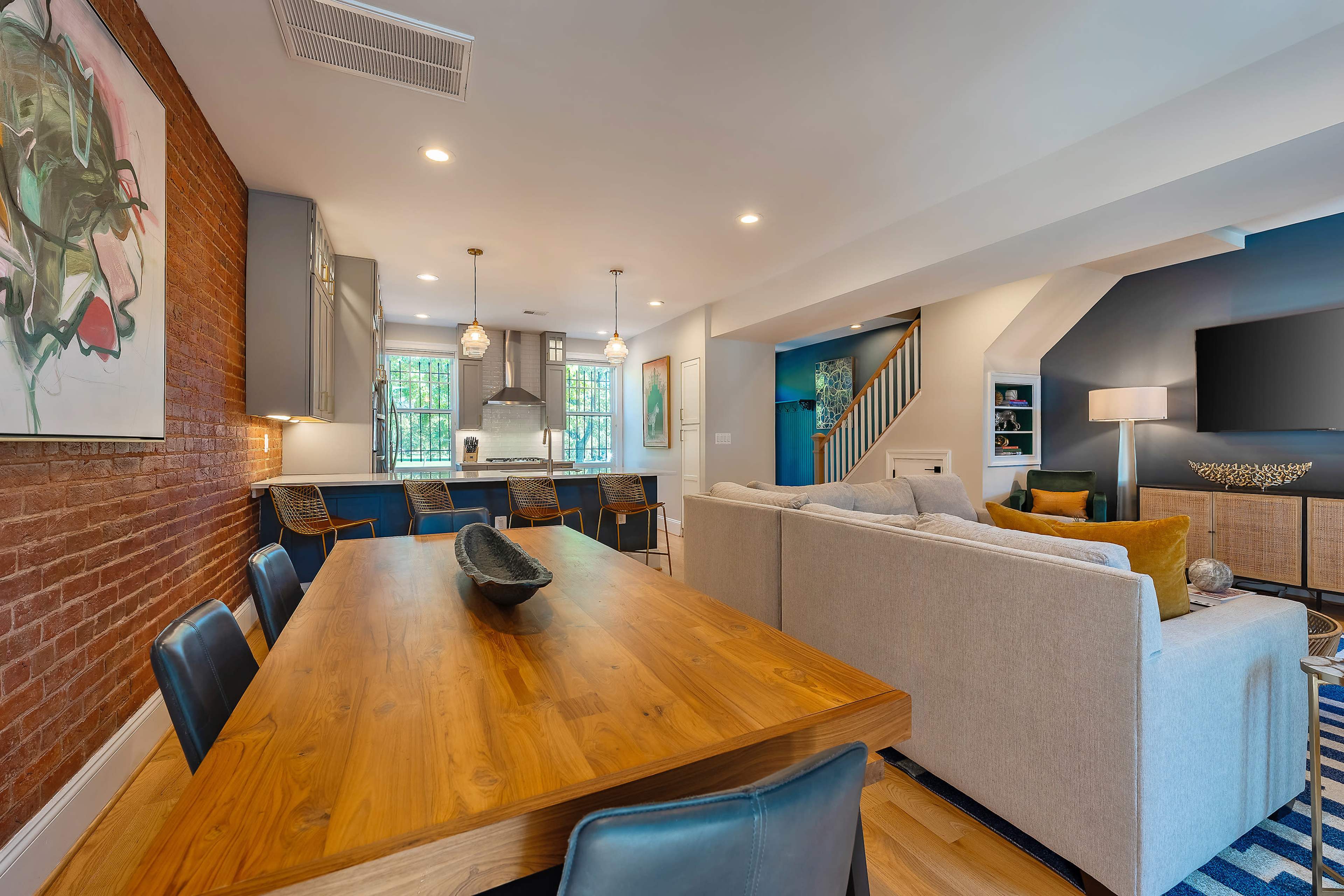 A modern open-concept living and dining area with a wooden dining table, a light-colored sofa, and a kitchen in the background featuring a large island and bar stools.