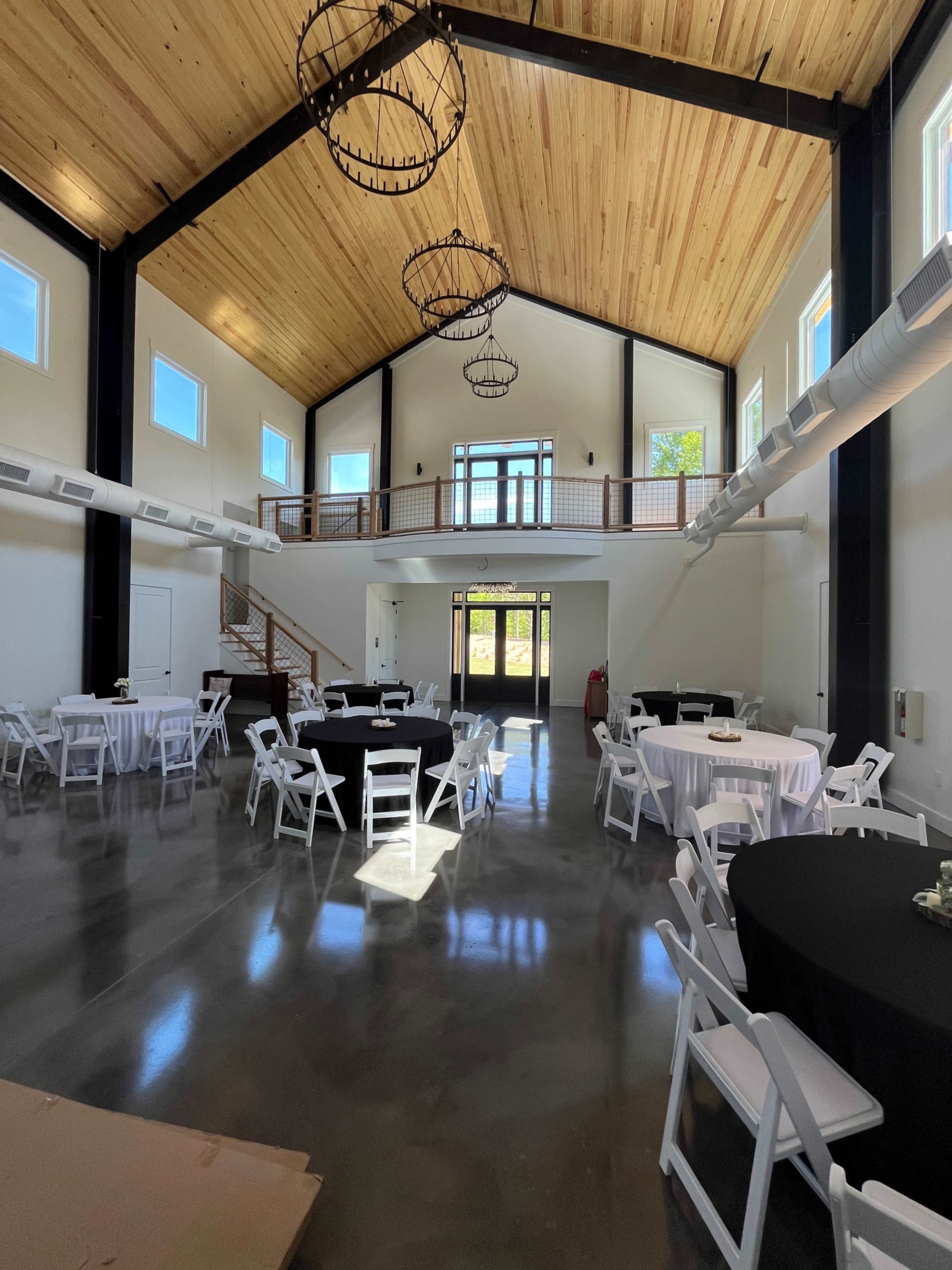 A spacious event hall with high ceilings, wooden beams, several round and rectangular tables, and a staircase leading to an upper level.