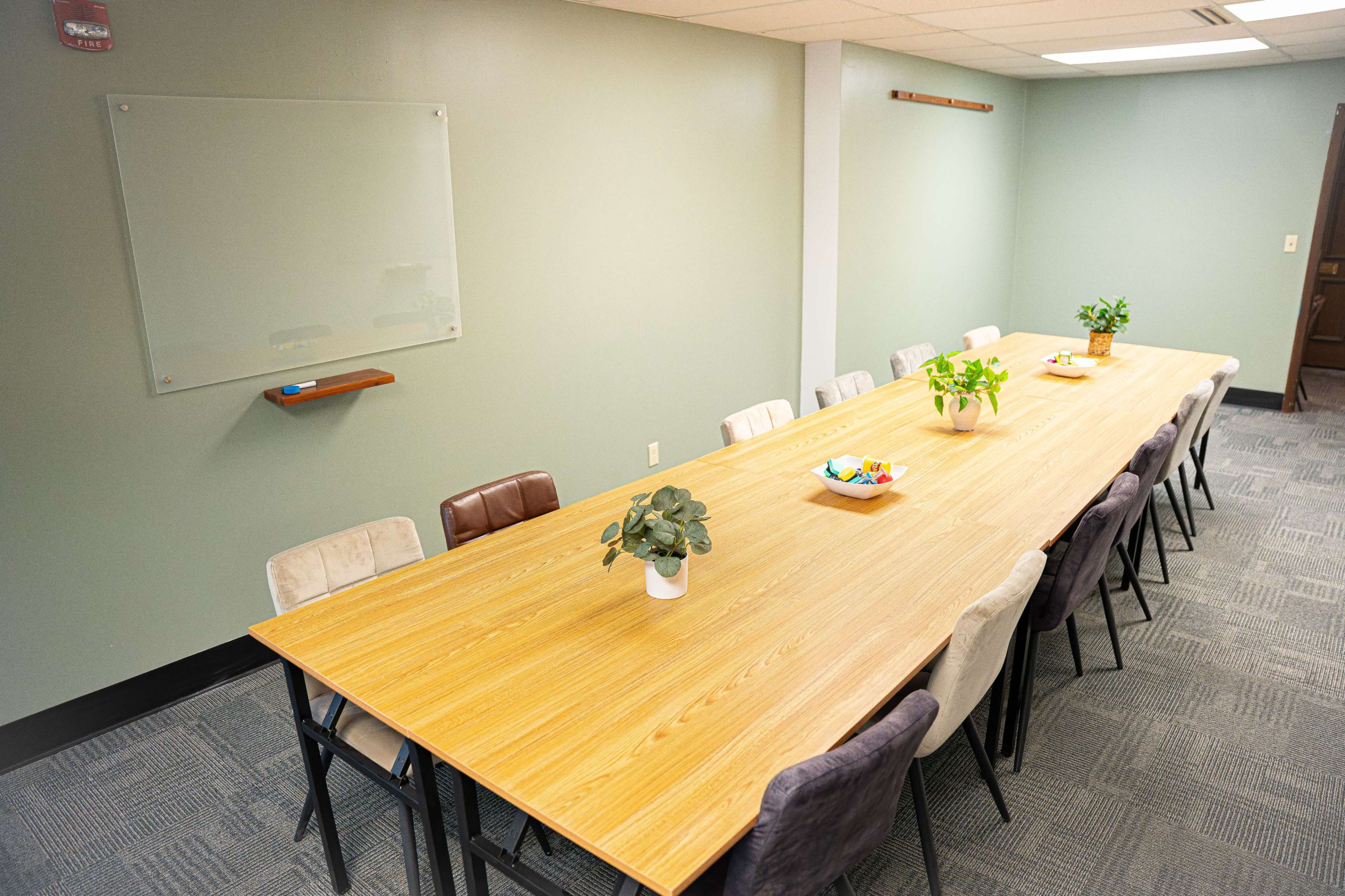 The image shows a spacious conference room with a long wooden table surrounded by chairs and two potted plants at opposite ends.