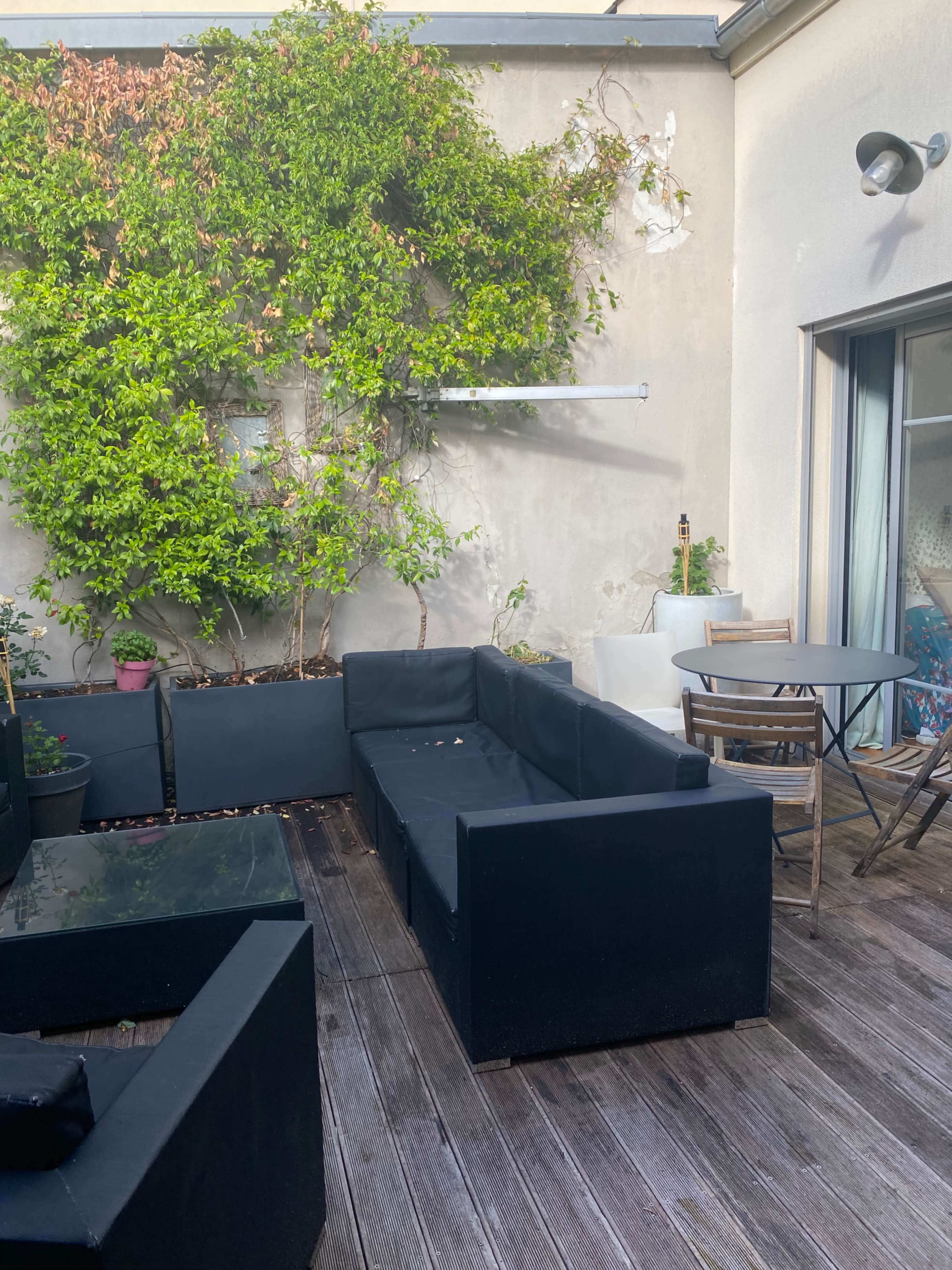 A patio area features black outdoor furniture arranged around a wooden deck, with potted plants and a small table in the background.