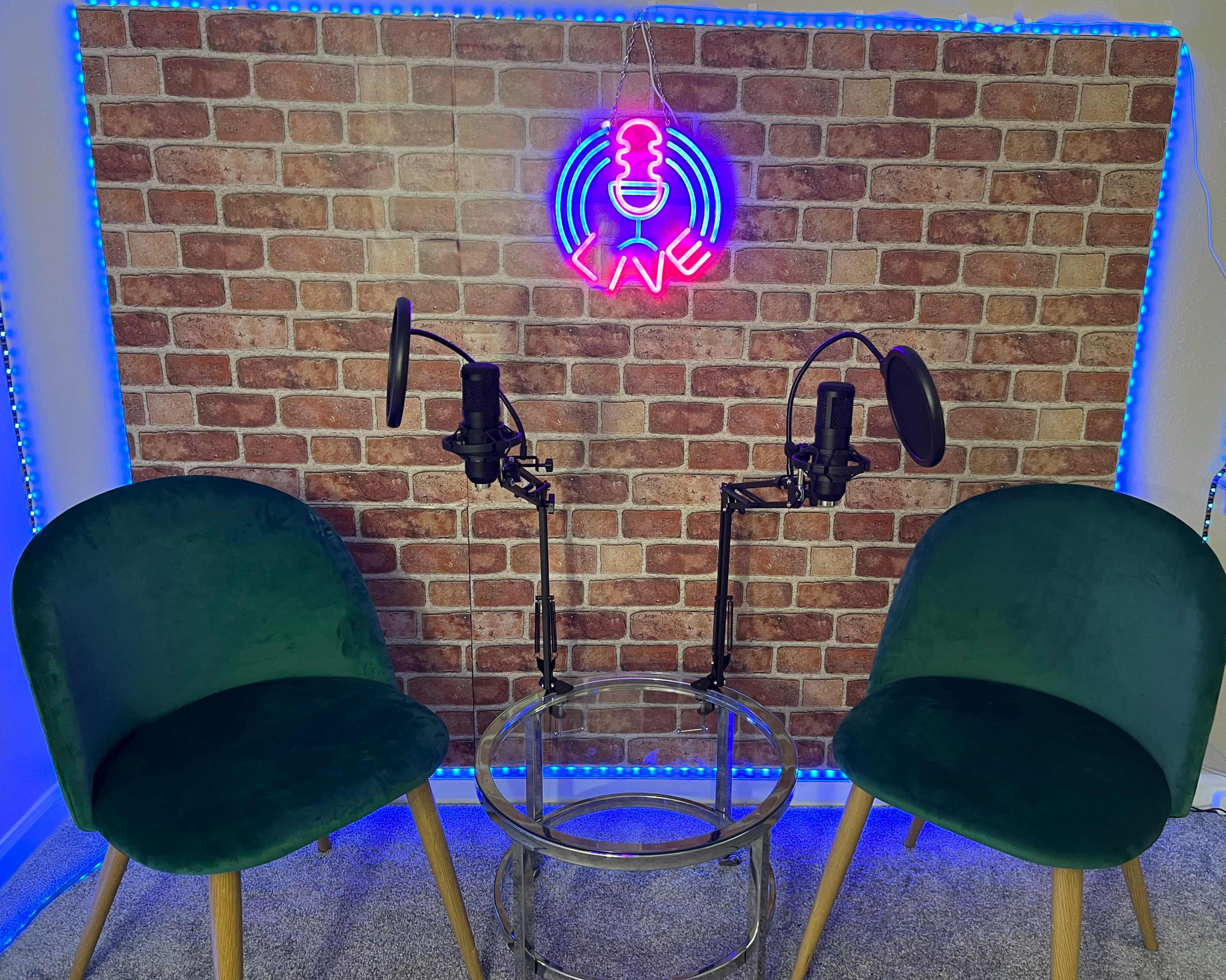 The image shows two green chairs positioned on either side of a glass table, with two microphones mounted on stands in front of a brick-patterned backdrop illuminated by blue lights and a neon sign.