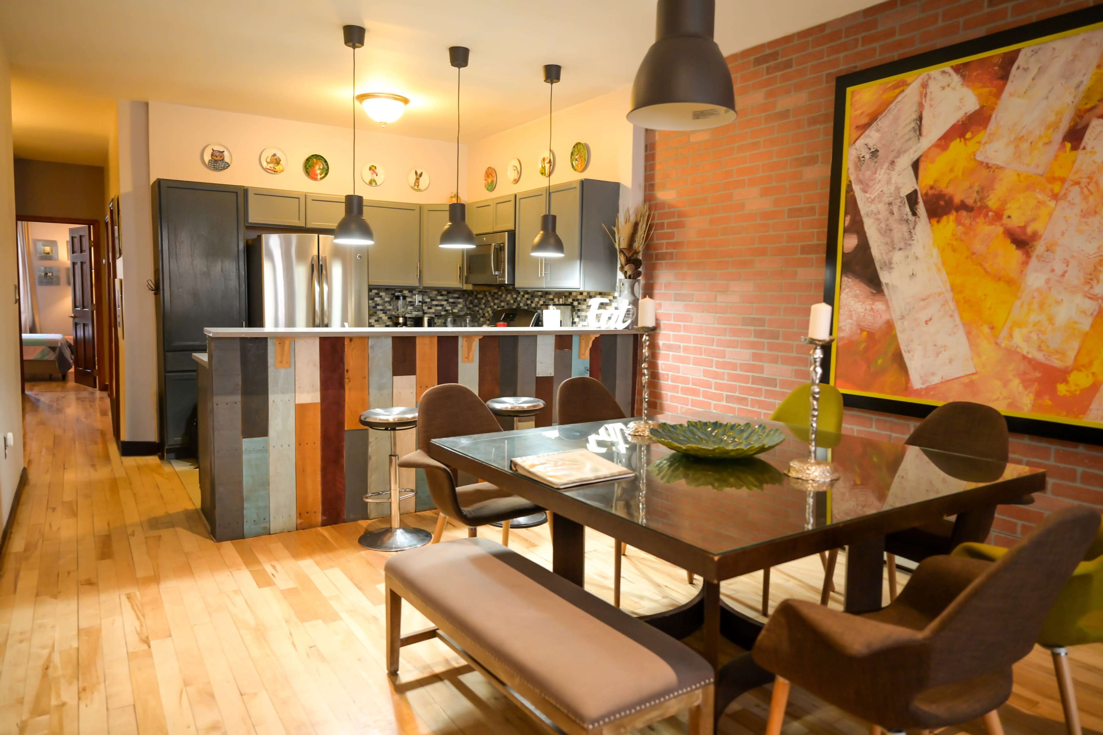 A modern kitchen and dining area features a wooden table with upholstered chairs, a bar with a colorful panel design, and exposed brick walls.