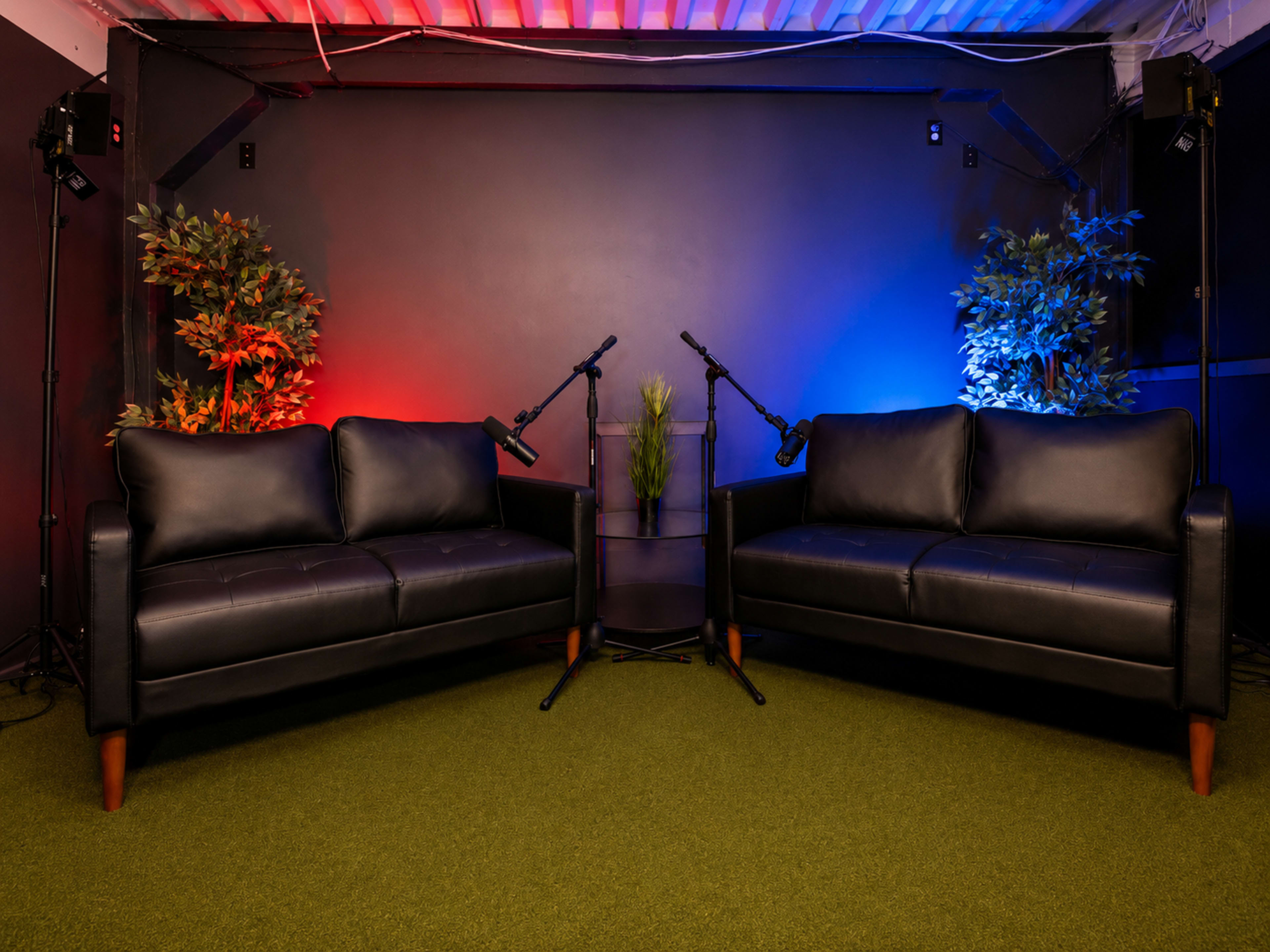 The scene features two black leather sofas facing each other on a green carpet, with a small table in the center and two microphones positioned nearby, illuminated by red and blue lighting.