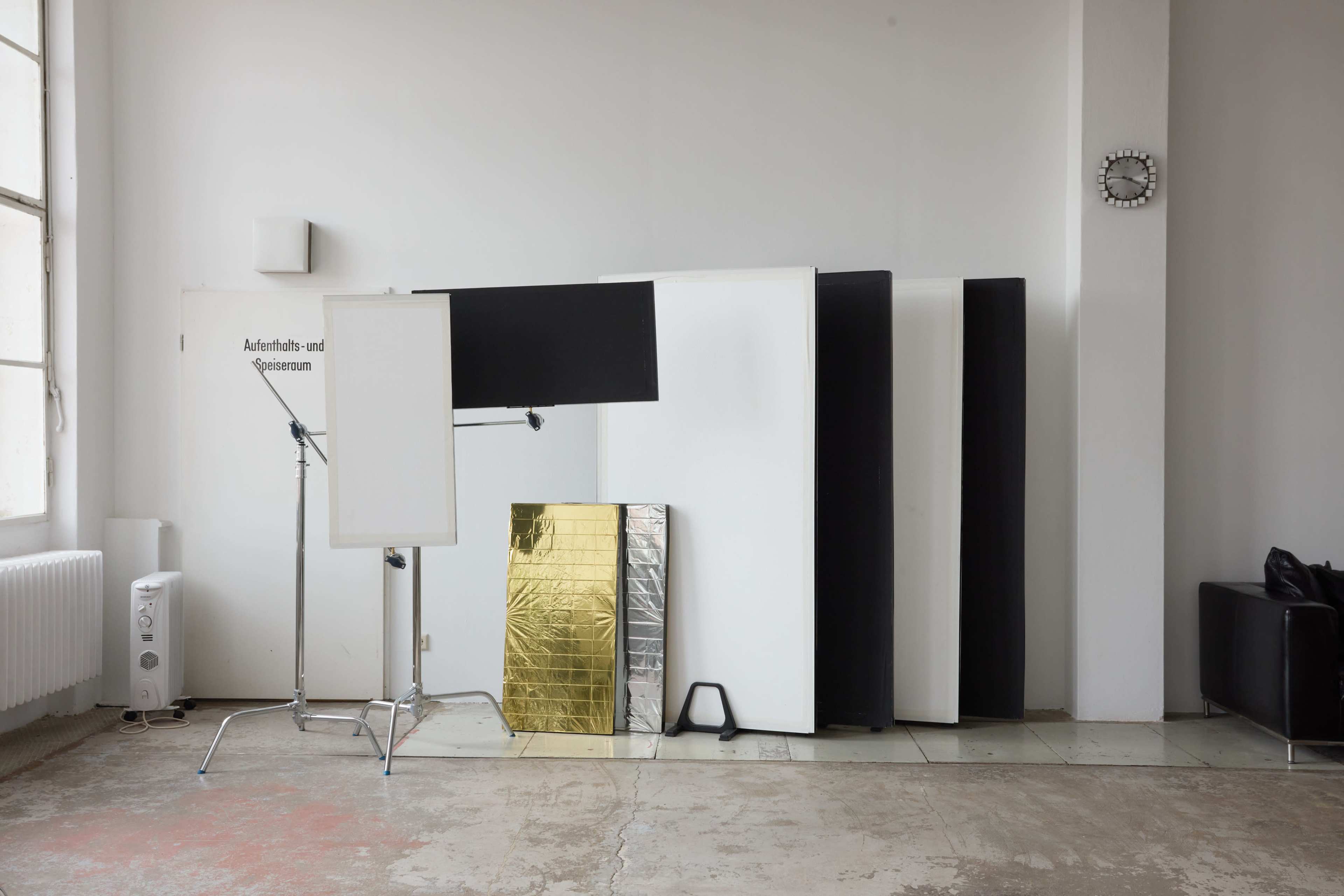 The image shows a minimalist studio space with various panels, a reflective board, and a standing frame arranged along a wall.
