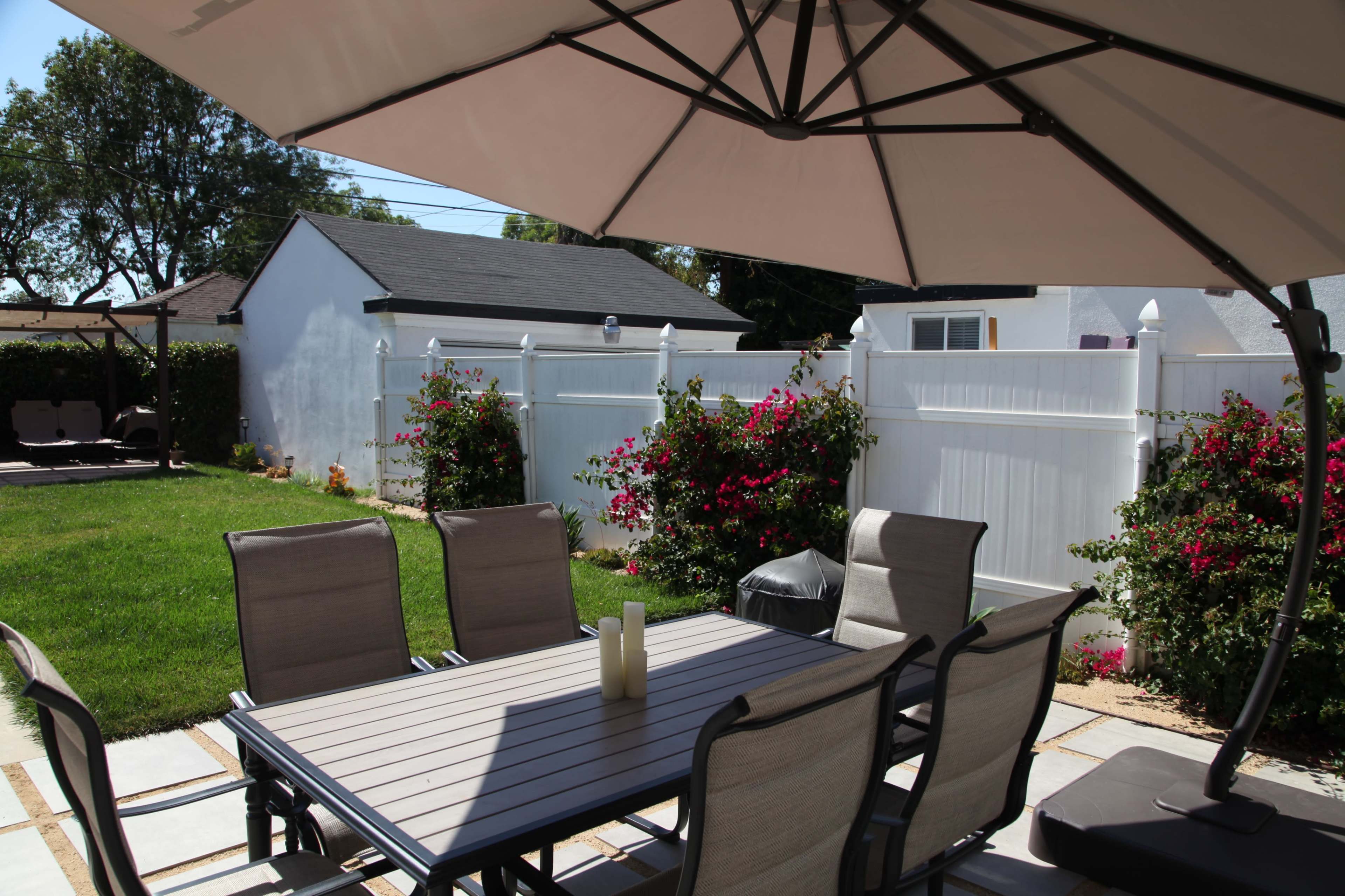 A patio area features a table with chairs under a large umbrella, surrounded by a well-maintained lawn and flowering bushes against a white fence.