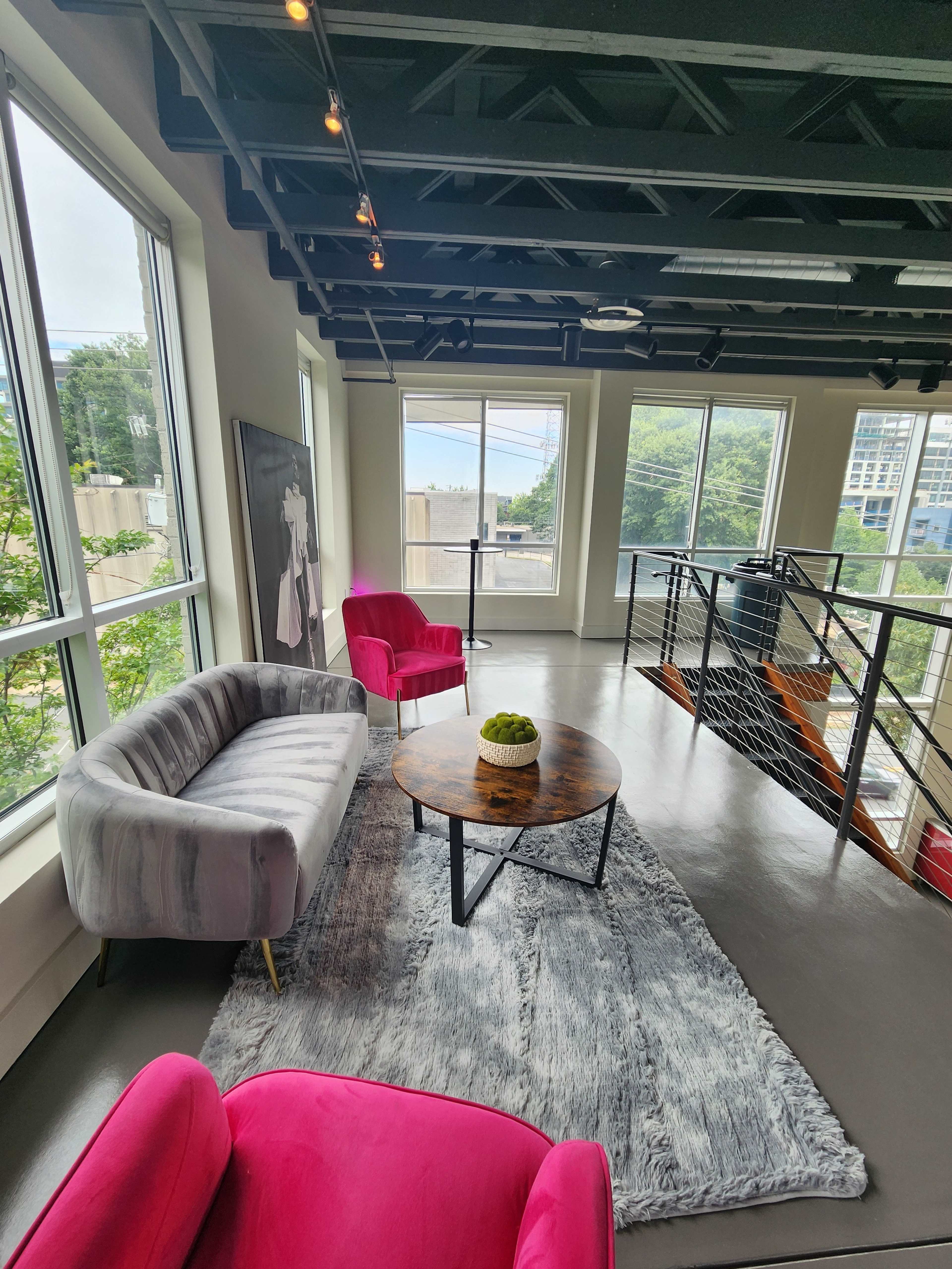 A modern lounge area with a gray sofa, two pink armchairs, a round coffee table, and large windows allowing natural light to enter.