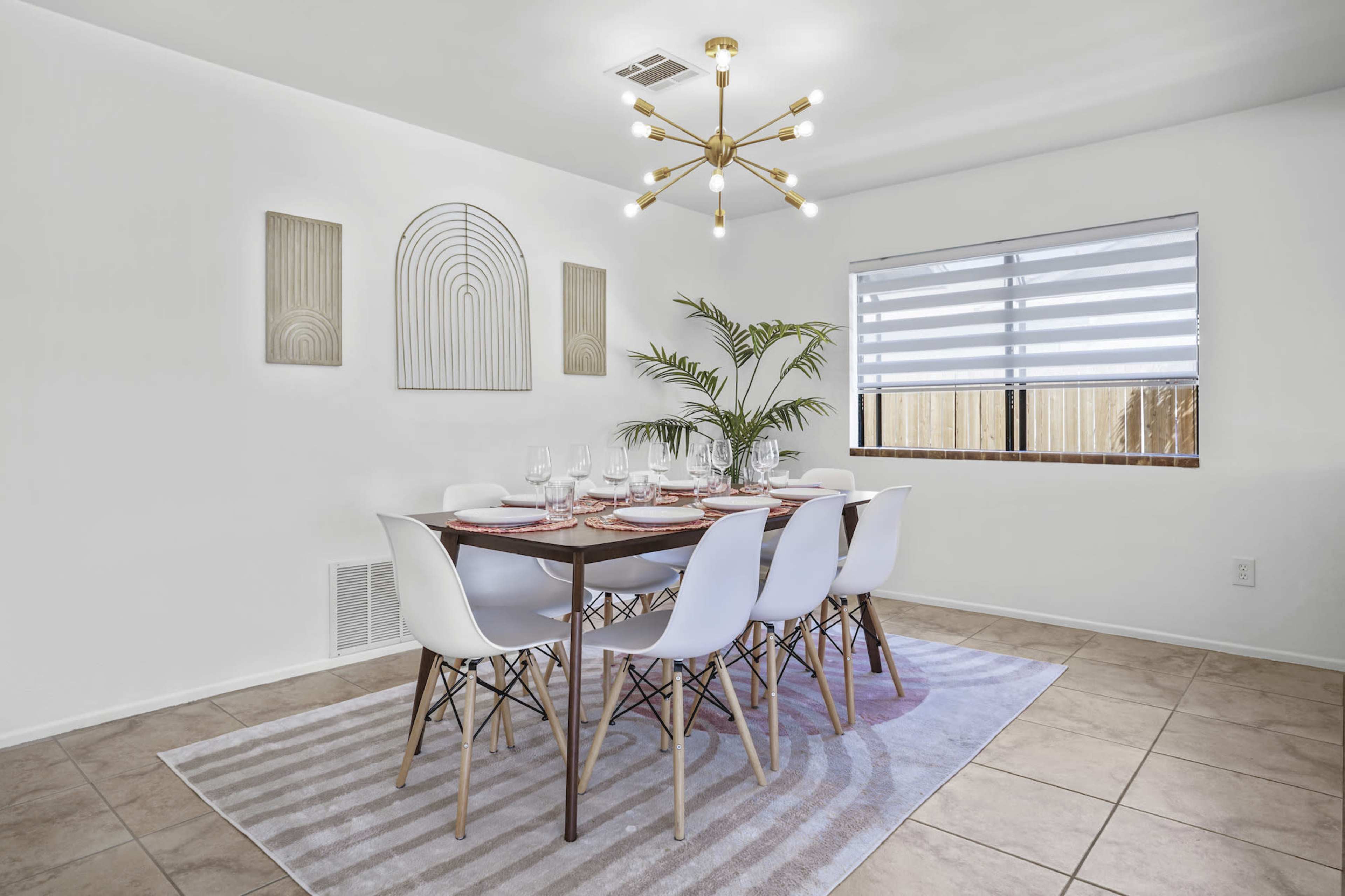 A modern dining room features a large wooden table set for dinner, surrounded by white chairs, with an overhead light fixture and decorative wall art.